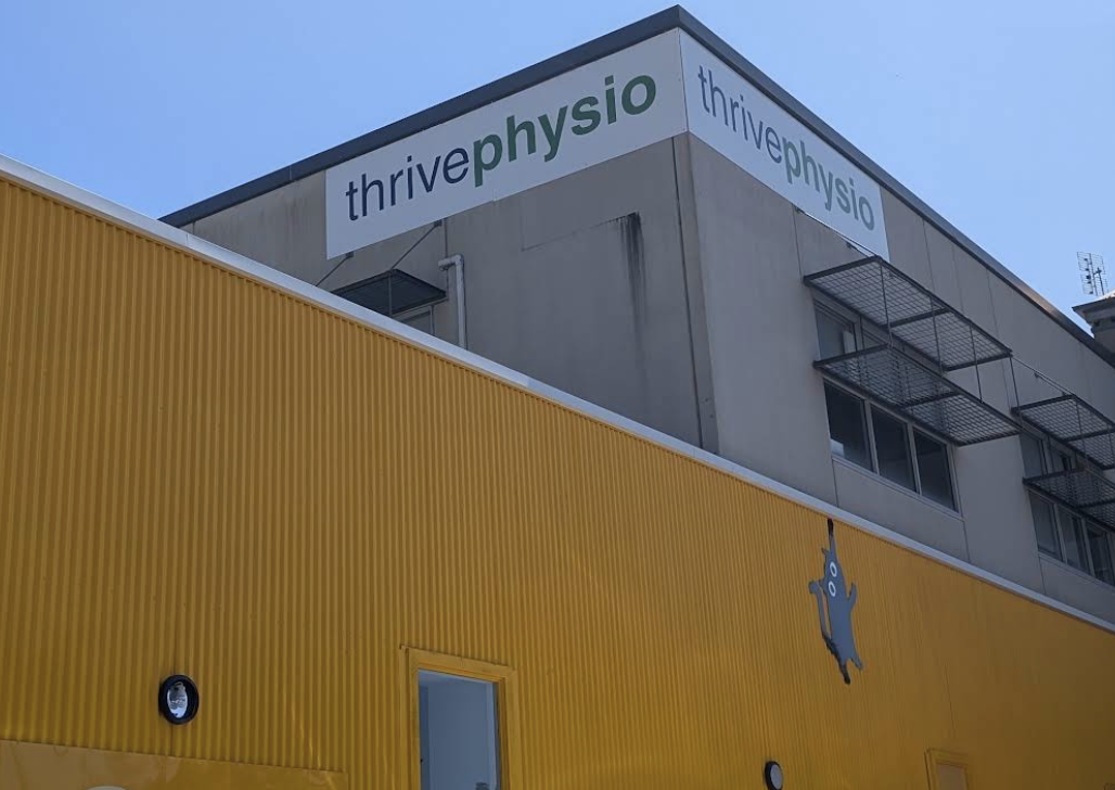 The clinic location with a sign reading 'thrive physio' on top and a yellow facade with a hanging decorative figur. The outside location of the physio clinic.