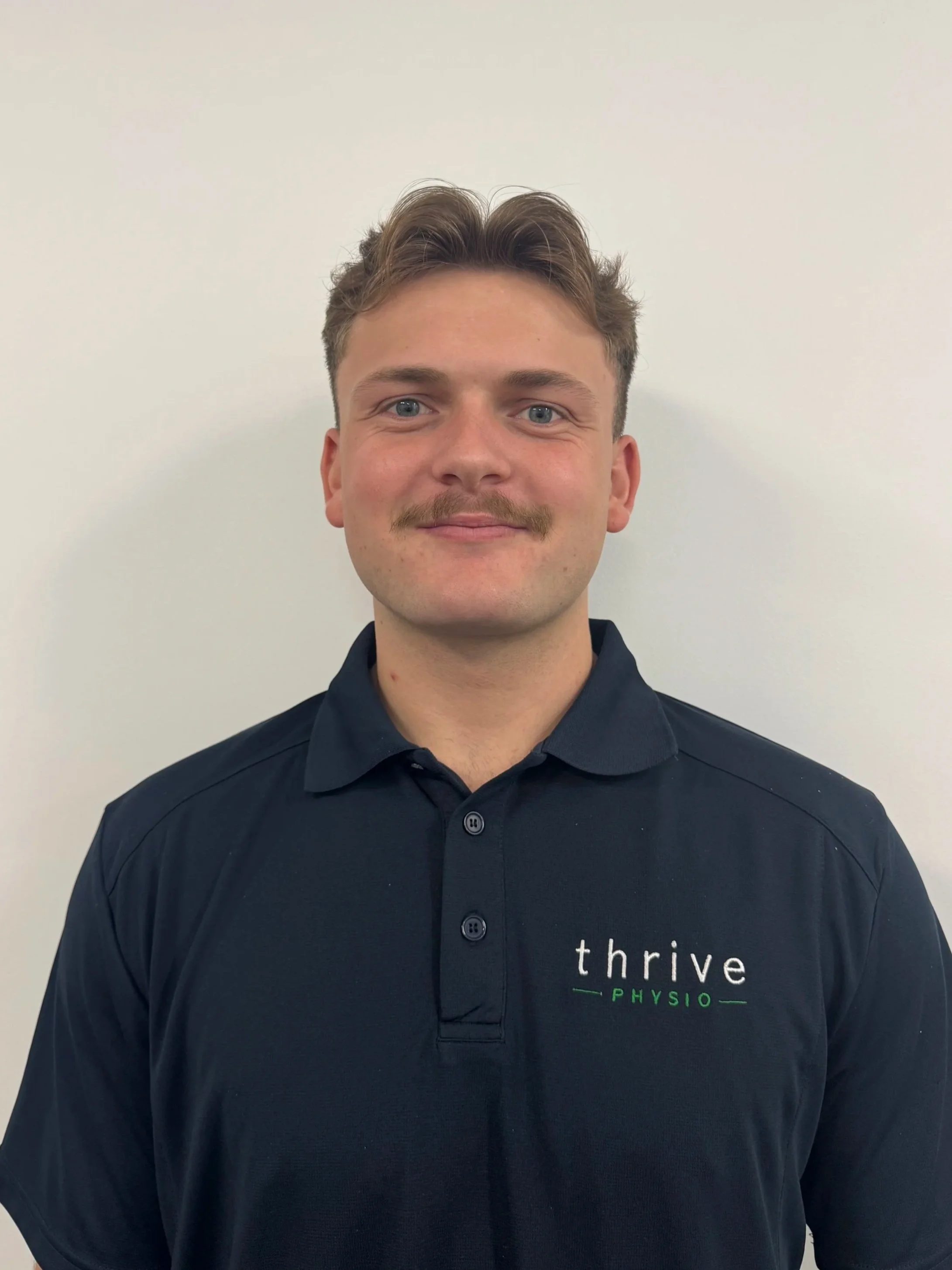 A young man with short light brown hair, blue eyes, and a mustache, smiling, wearing a black polo shirt with 'thrive physi' logo on the chest, standing against a plain white wall.