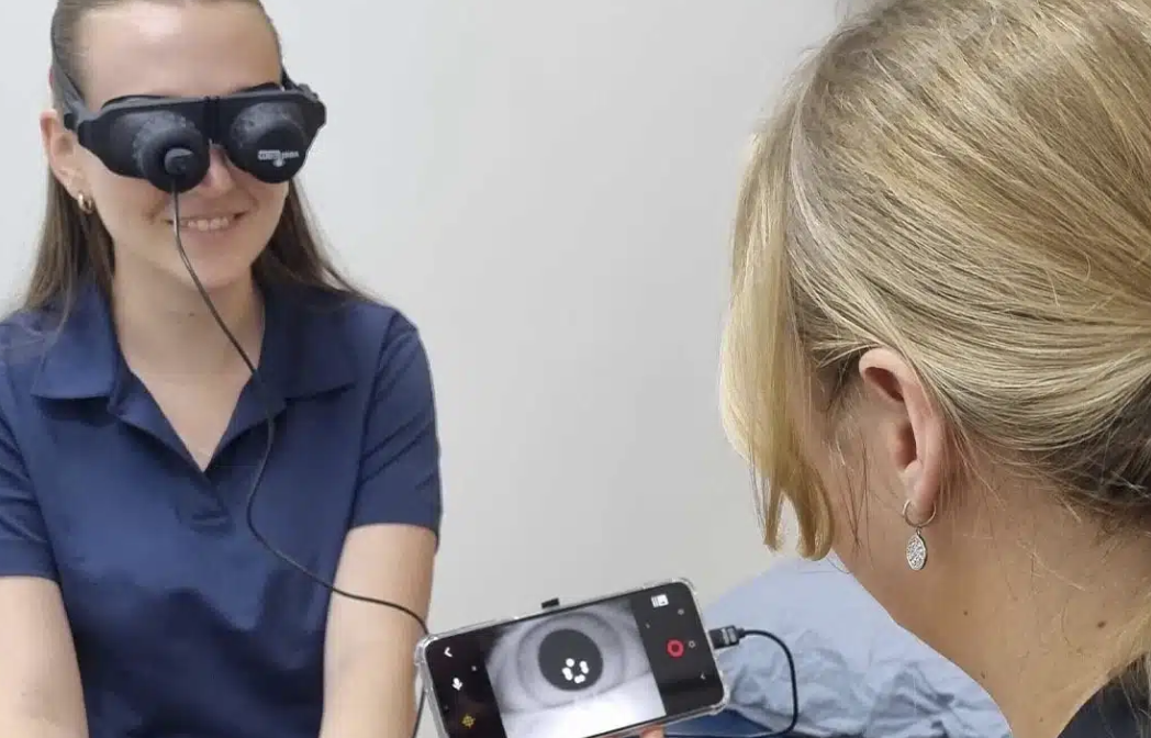 A vestibular physiotherapy appointment in a clinical setting; one woman wearing vestibular goggles and the other woman looking at a phone screen showing a camera view.