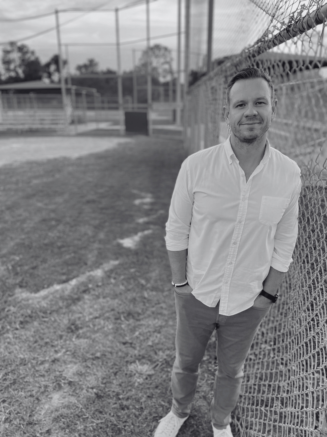 A man standing outdoors beside a chain-link fence, wearing a white shirt and light-colored pants, with hands in pockets and smiling.