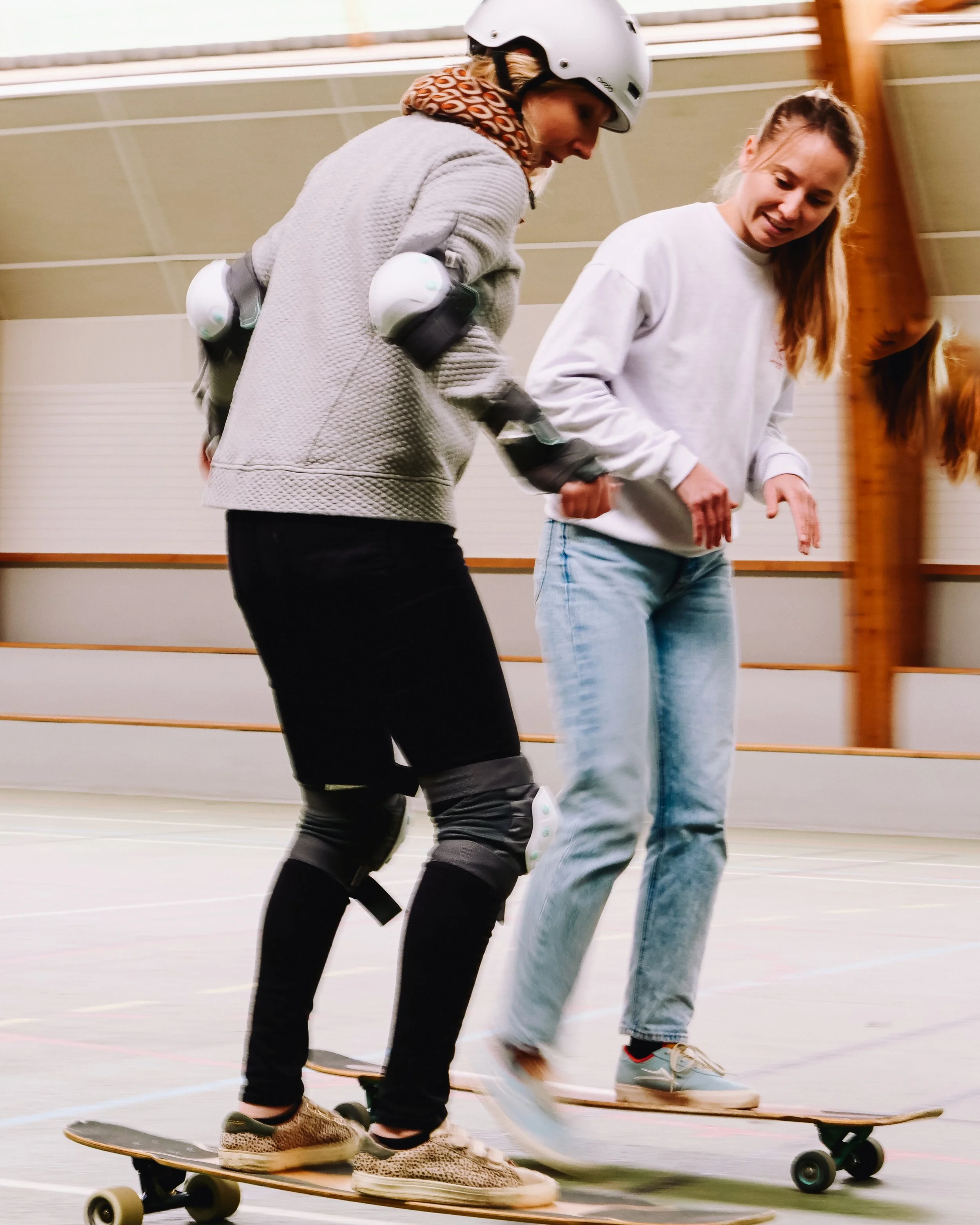 Deux jeunes femmes apprennent à faire du skateboard dans une salle. La femme de gauche porte un casque, des protections et un sweat gris, tandis que la femme de droite porte un sweat blanc et des jeans.