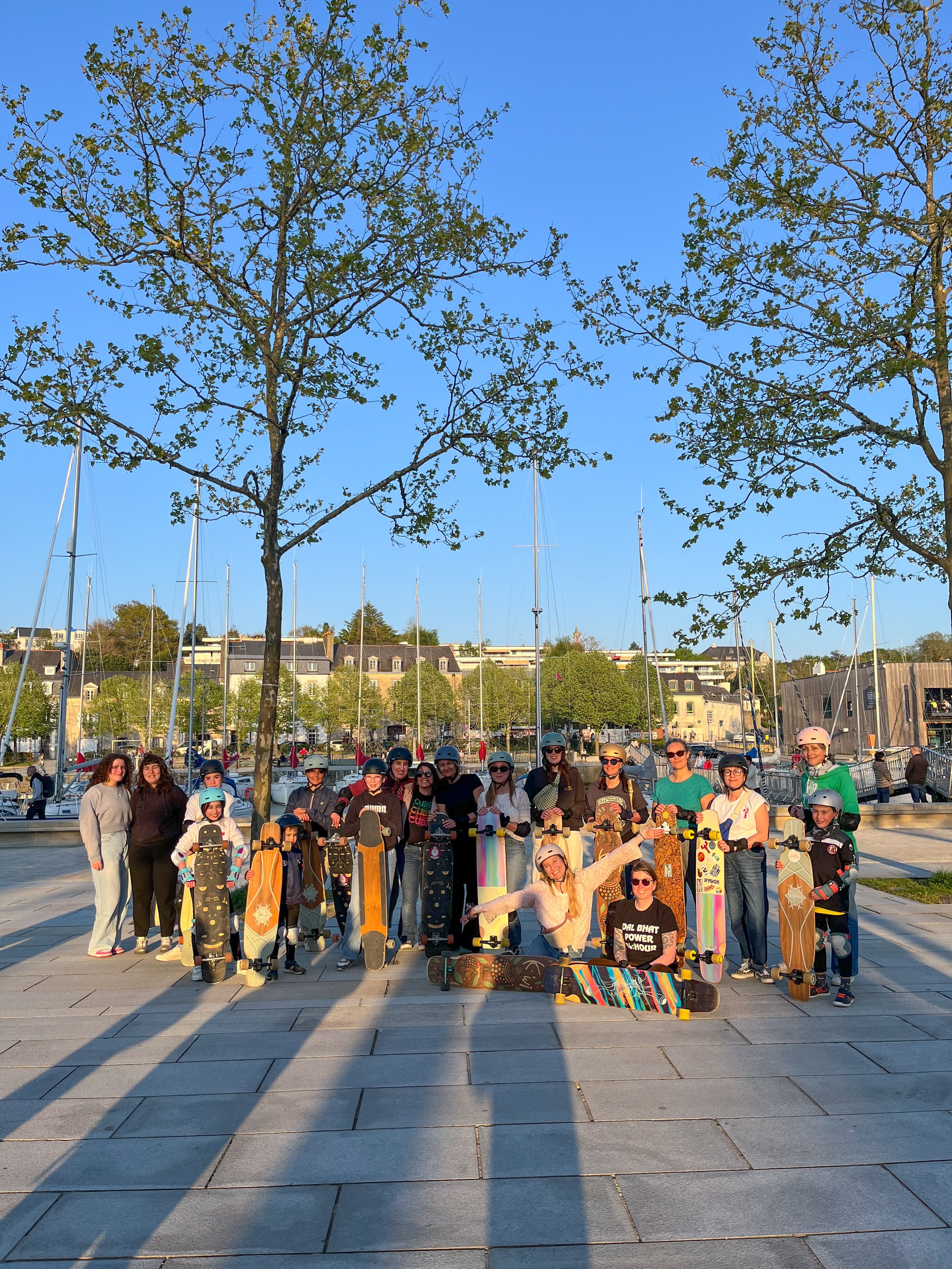Groupe de personnes de différents âges avec des skateboards et des casques, posant pour une photo en plein air près du port avec des voiliers et des bâtiments Au fond, deux arbres et un ciel clair. Il semble qu'ils font partie d'une activité de groupe de skate.