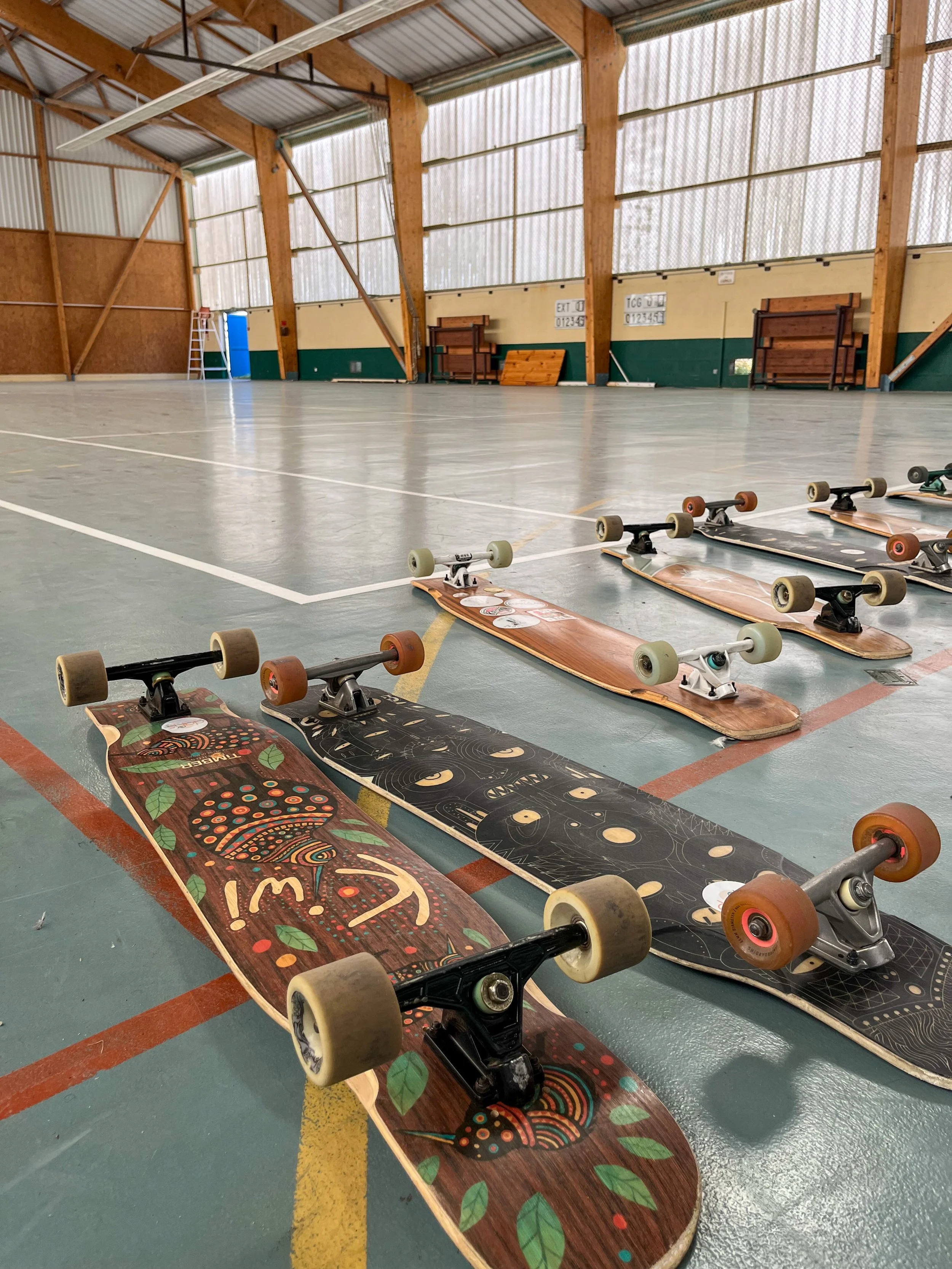 Des skateboards disposés sur le sol d'une salle de sport. La salle a une structure en bois et des murs hauts, avec des lignes marquées au sol et des équipements de sport rangés en arrière-plan.