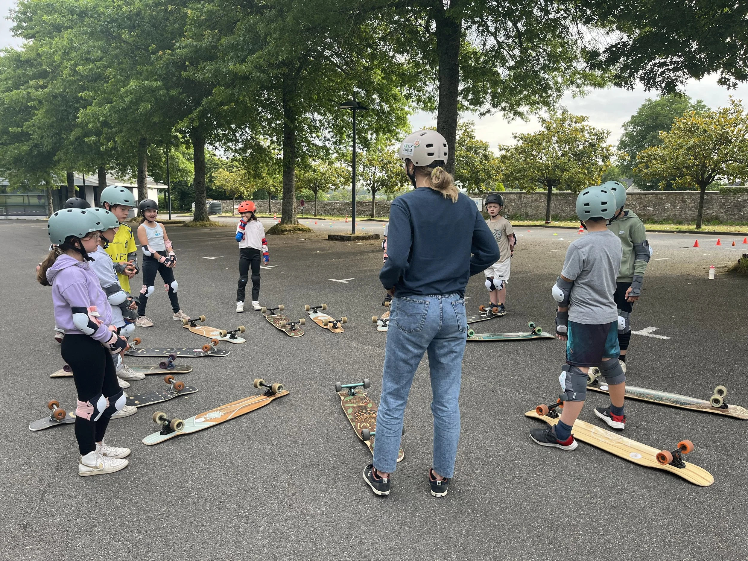Groupe d'enfants portant des casques et des protections, sur des longboards posés au sol, lors d'une session d'apprentissage du longboard dancing en extérieur avec une éducatrice diplômée.