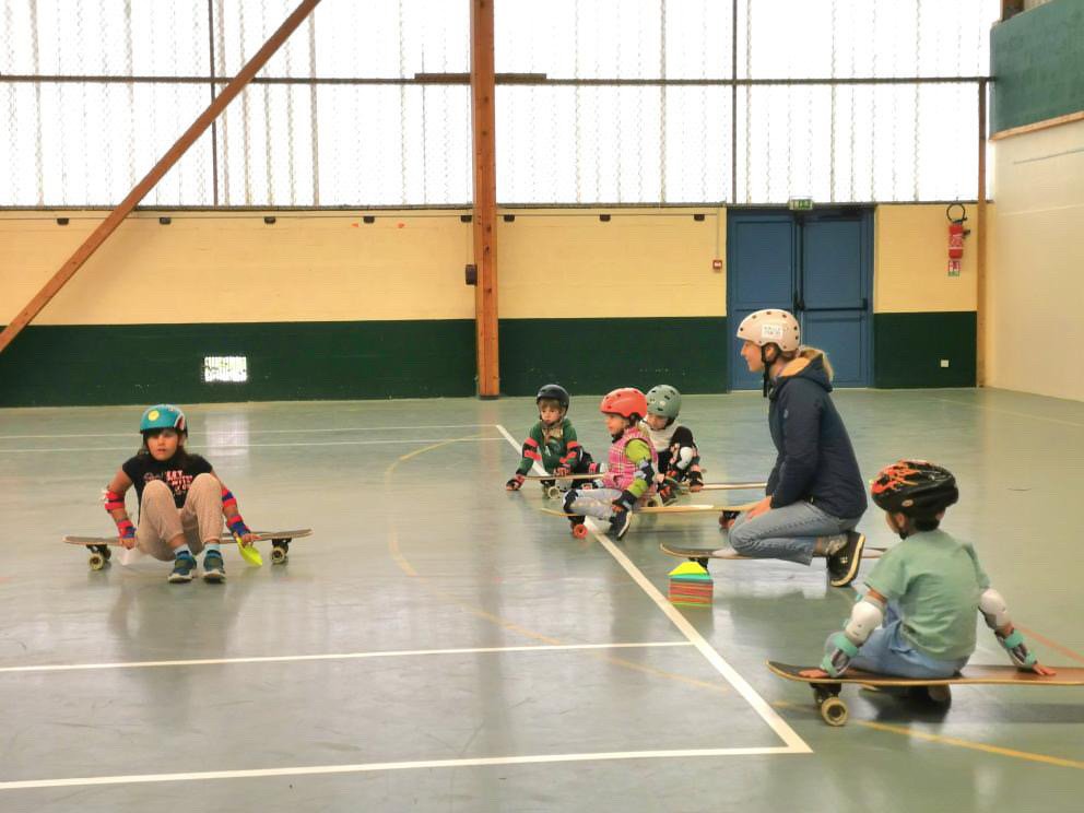 Entraînement de skate pour enfants dans une salle couverte, avec un instructeur et plusieurs enfants portant des casques et protections.