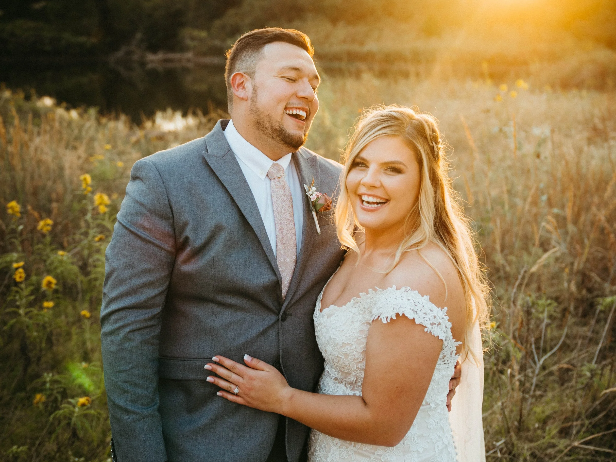A newlywed couple embracing outdoors during sunset, smiling happily, with a field of wildflowers and trees in the background.