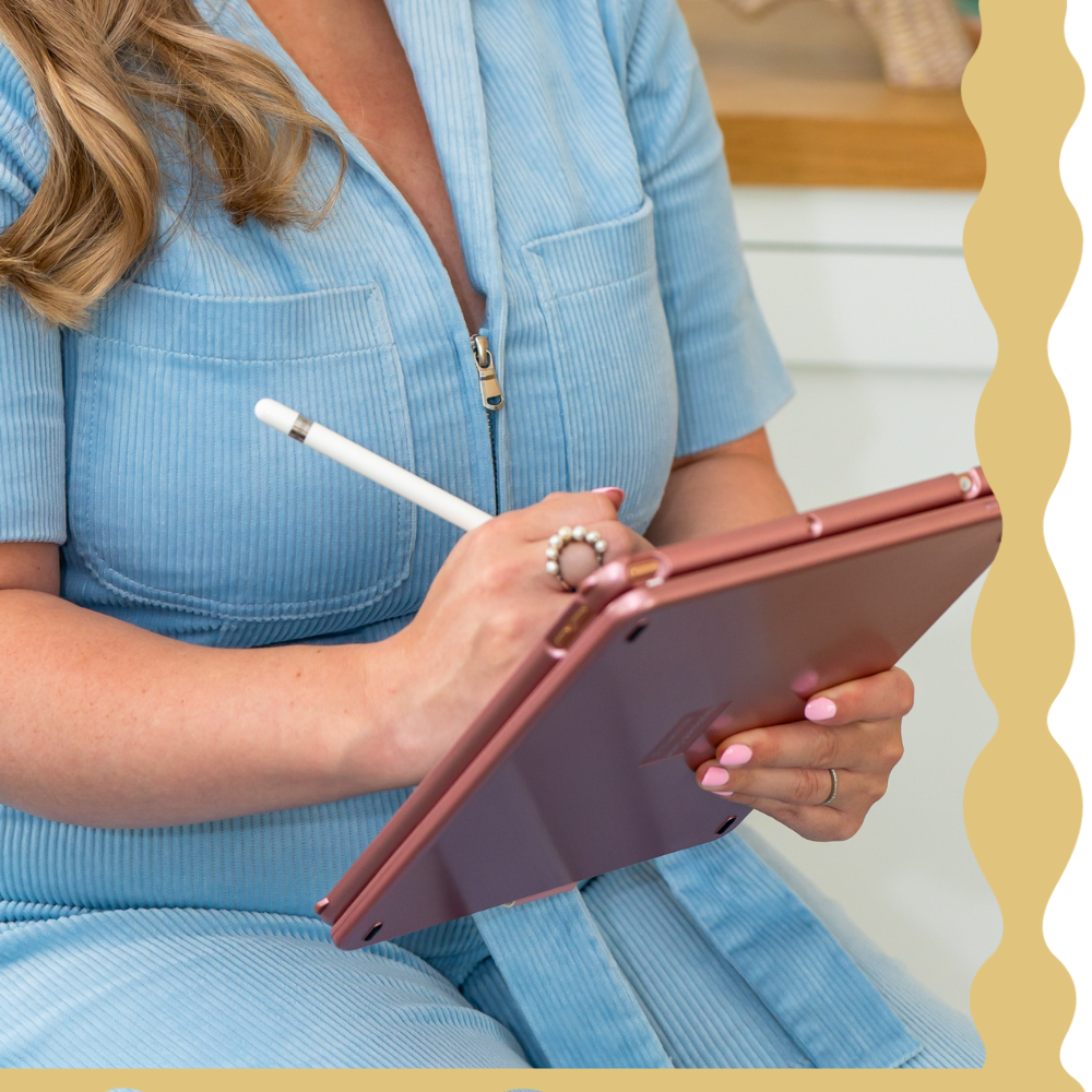 A woman wearing a light blue zipped top is sitting and writing on a digital tablet with a white stylus. She has pink painted nails and a pearl ring.
