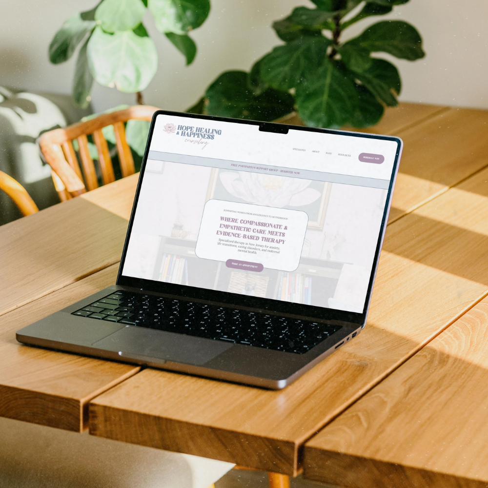 A laptop on a wooden table, displaying a website titled "Hope Healing & Happiness" with a focus on compassionate and empathetic care and evidence-based therapy. In the background, there are large green plants and a wooden chair.