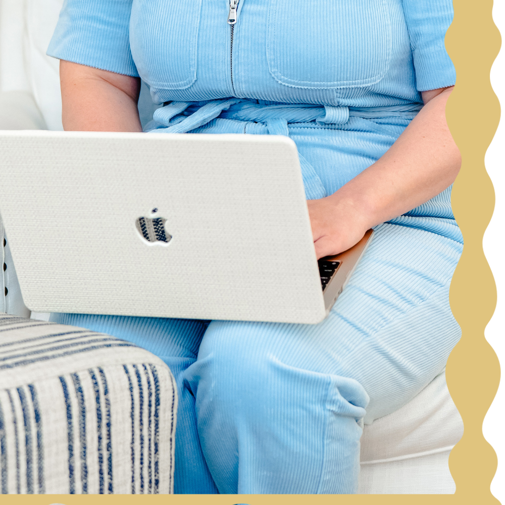 Person in blue pajamas sitting on a white sofa, using a silver laptop with an Apple logo.