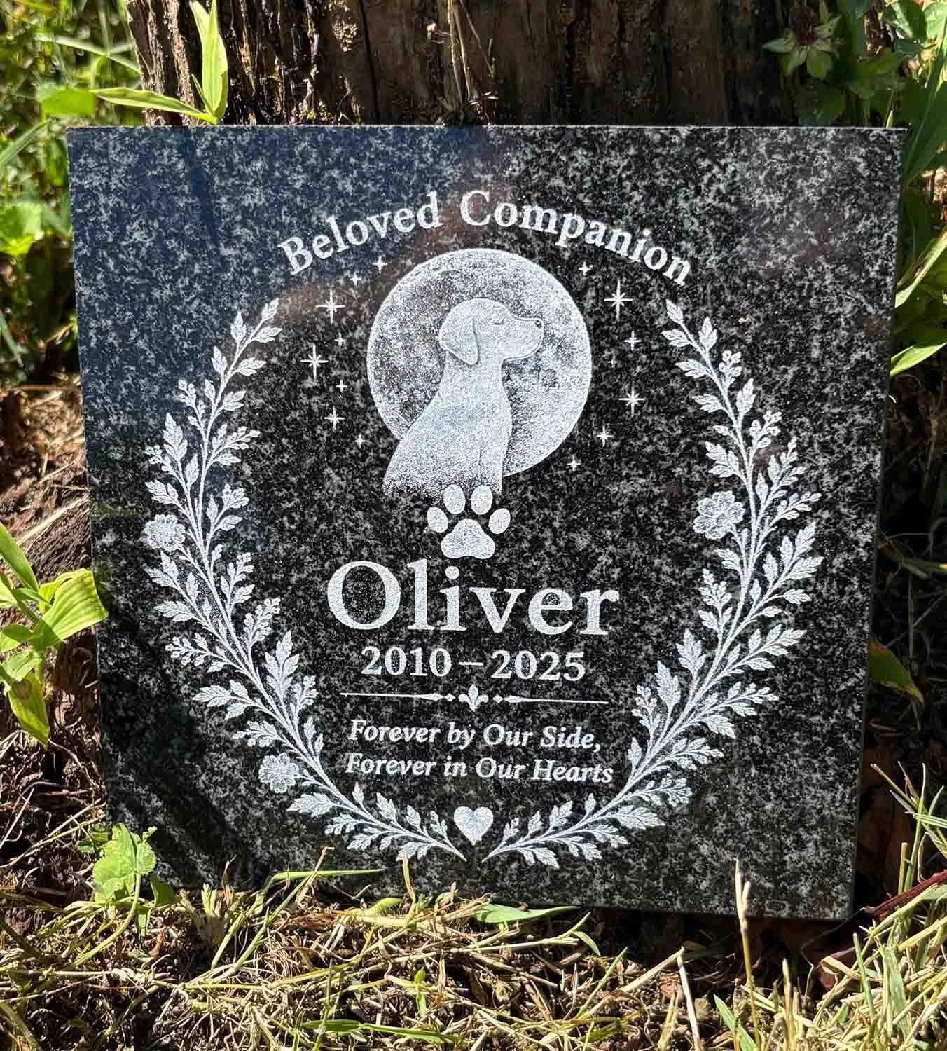 Personalized black granite dog grave marker with dog, moon, paw print and floral design displayed outdoors beside a tree.