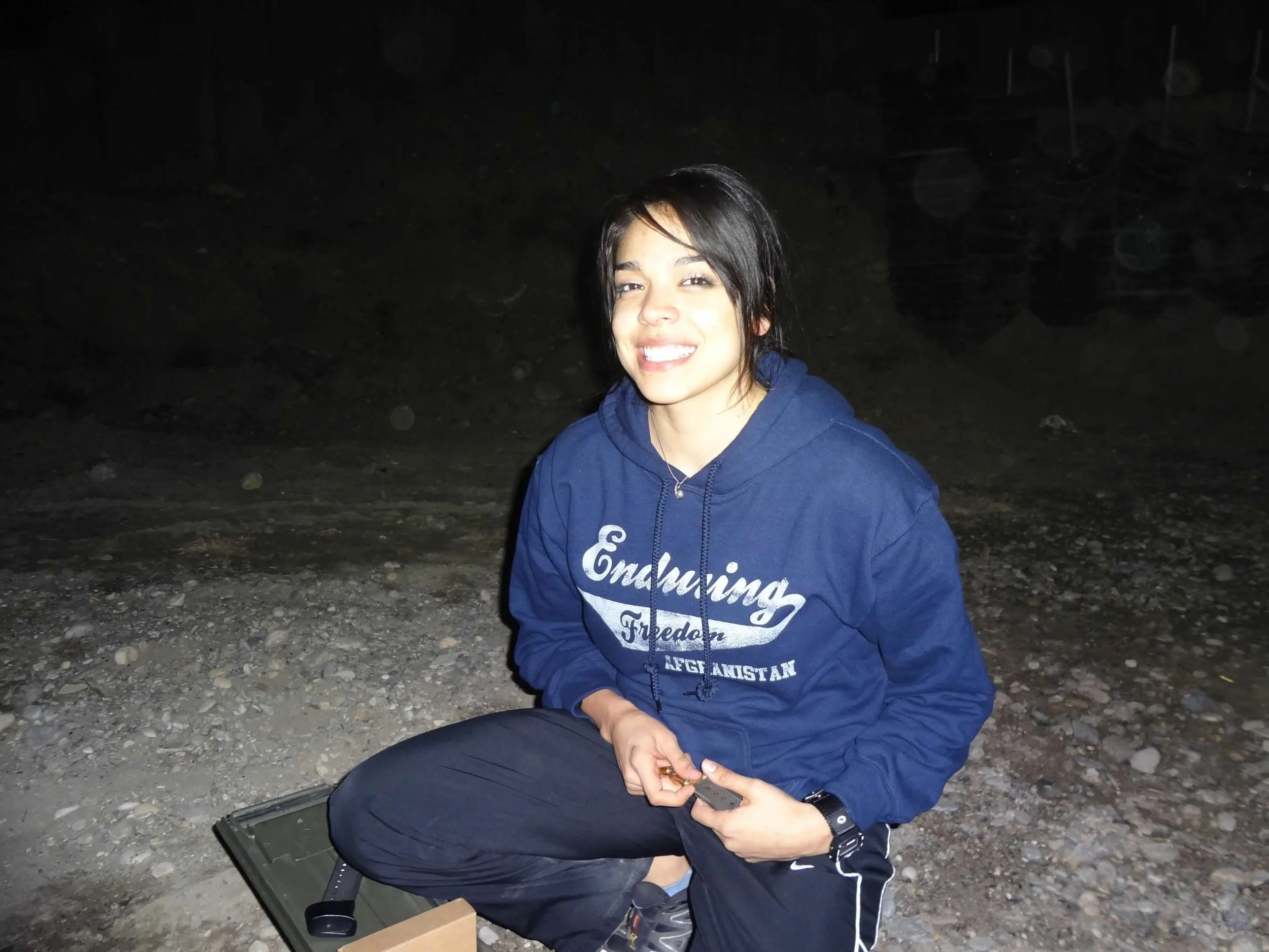 Smiling young woman with short dark hair sitting on rocks outdoors at night, wearing a blue hoodie with text and holding a small object in her hands.