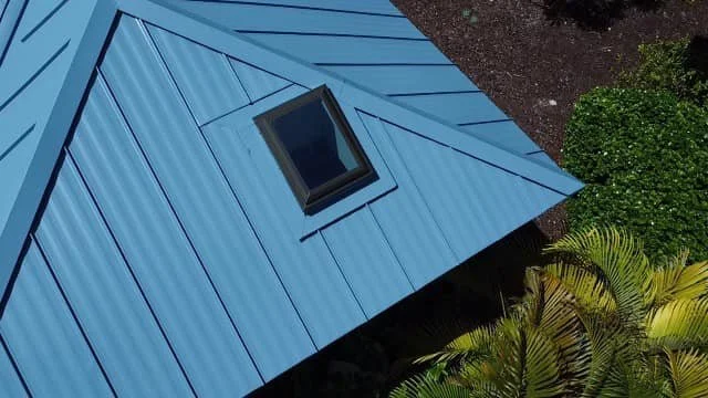 Blue metal roofing with a skylight, showcasing durability and modern design, surrounded by lush greenery.