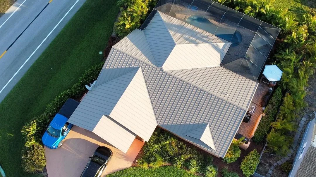 Aerial view of a residential home in Fort Myers, Florida, featuring a light-colored metal roof, surrounded by lush greenery, with parked vehicles in the driveway.