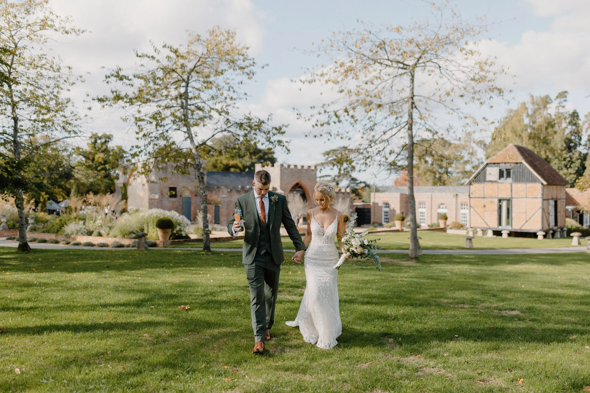Bride and groom walking on a lawn, holding hands, with rustic buildings in the background.