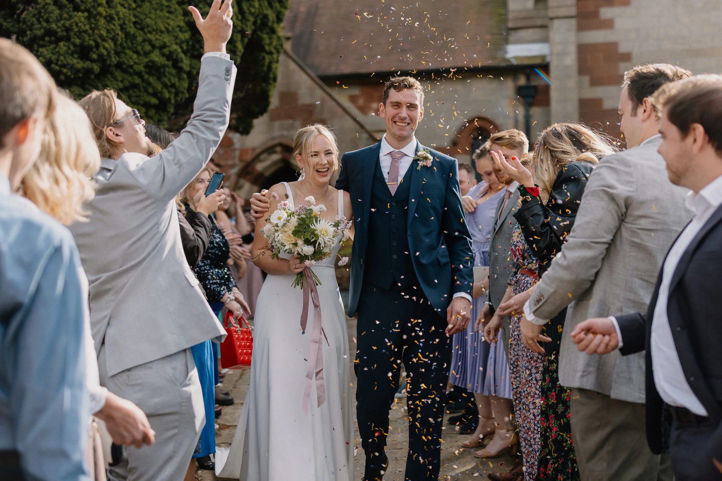 Bride and groom walking through a crowd celebrating, with confetti falling, outside a church.