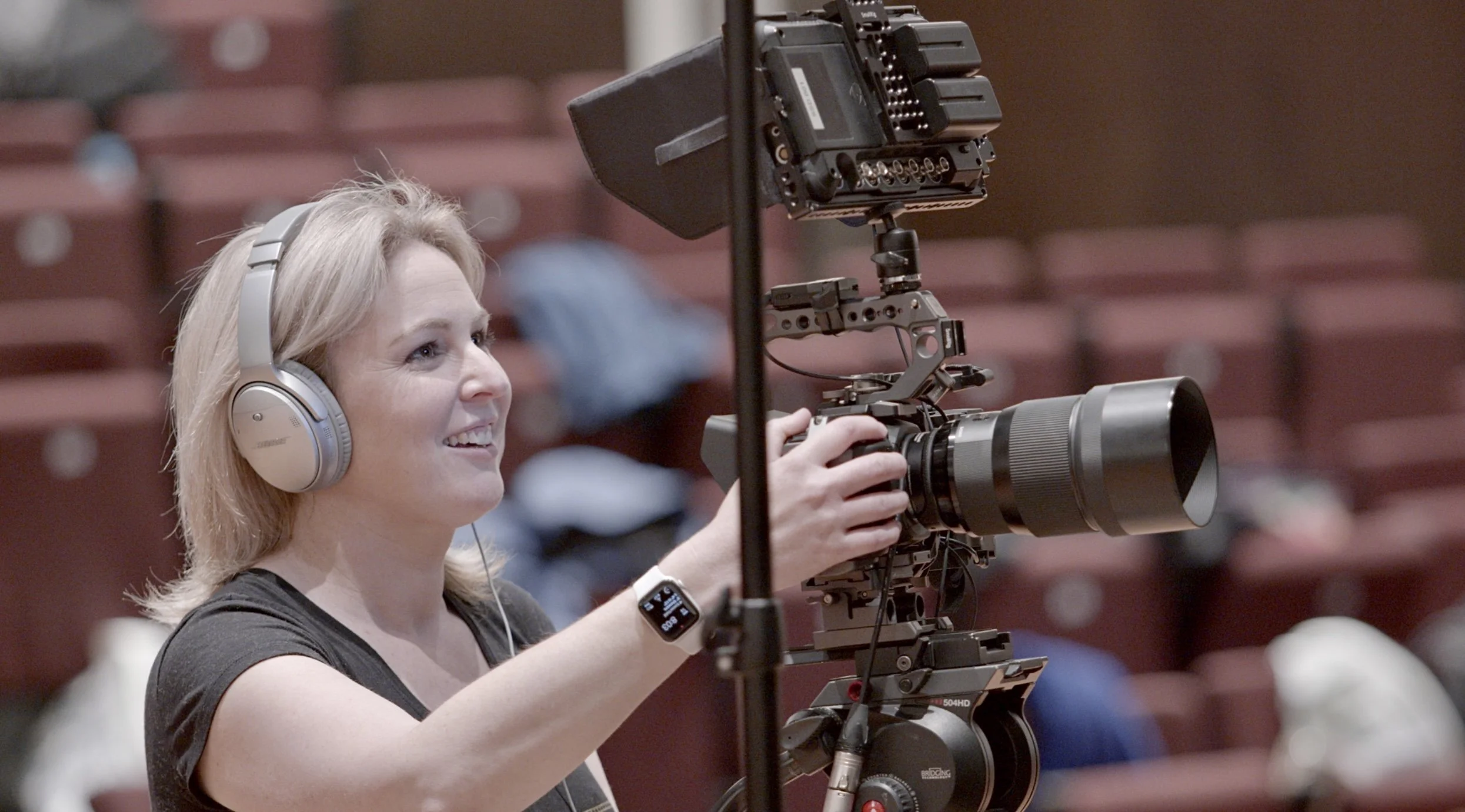 A woman with headphones operating a professional video camera in a theater or auditorium setting.