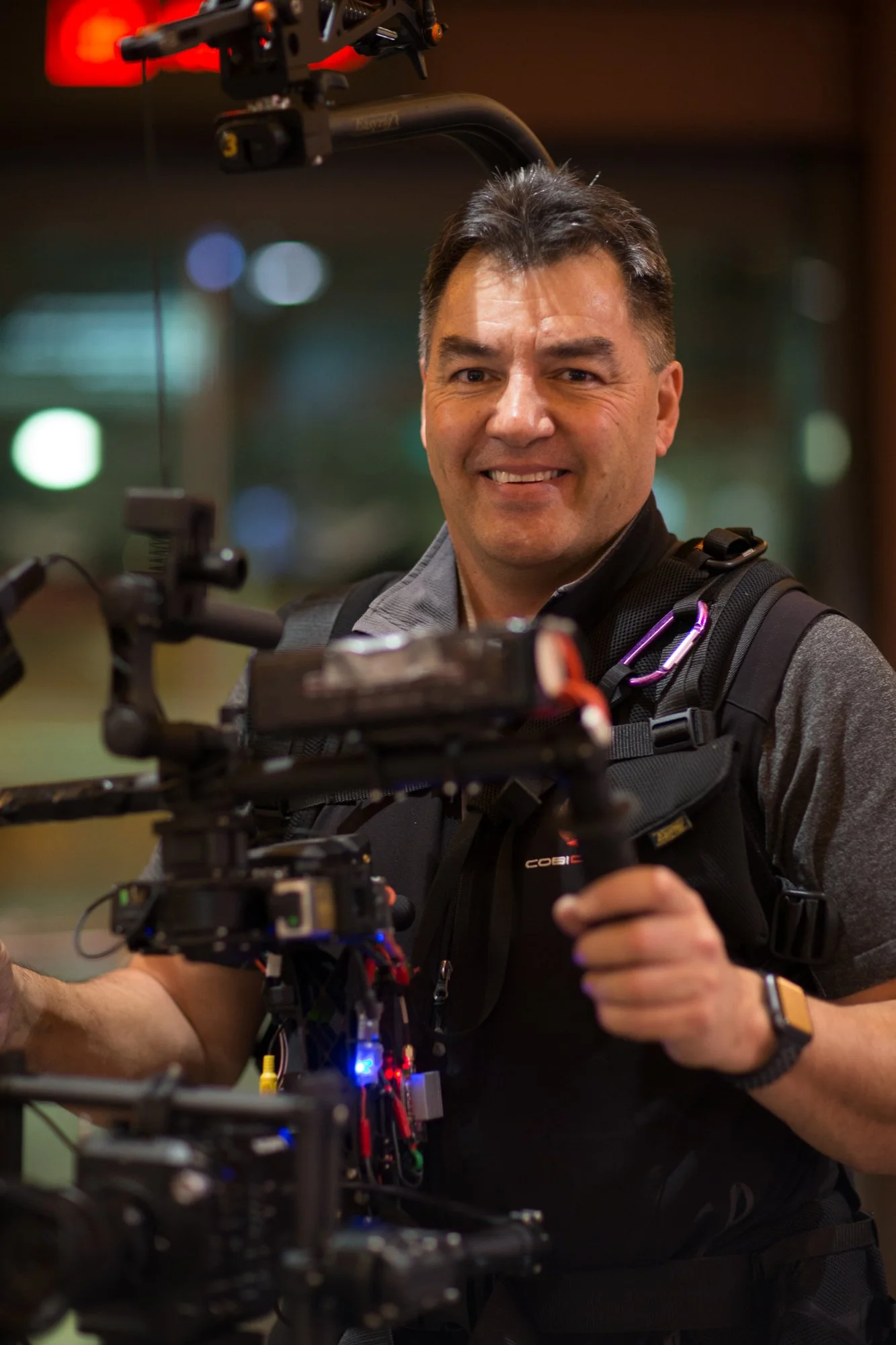 A smiling man operating a professional camera rig in an indoor setting, wearing a black vest, gray shirt, and a smartwatch.