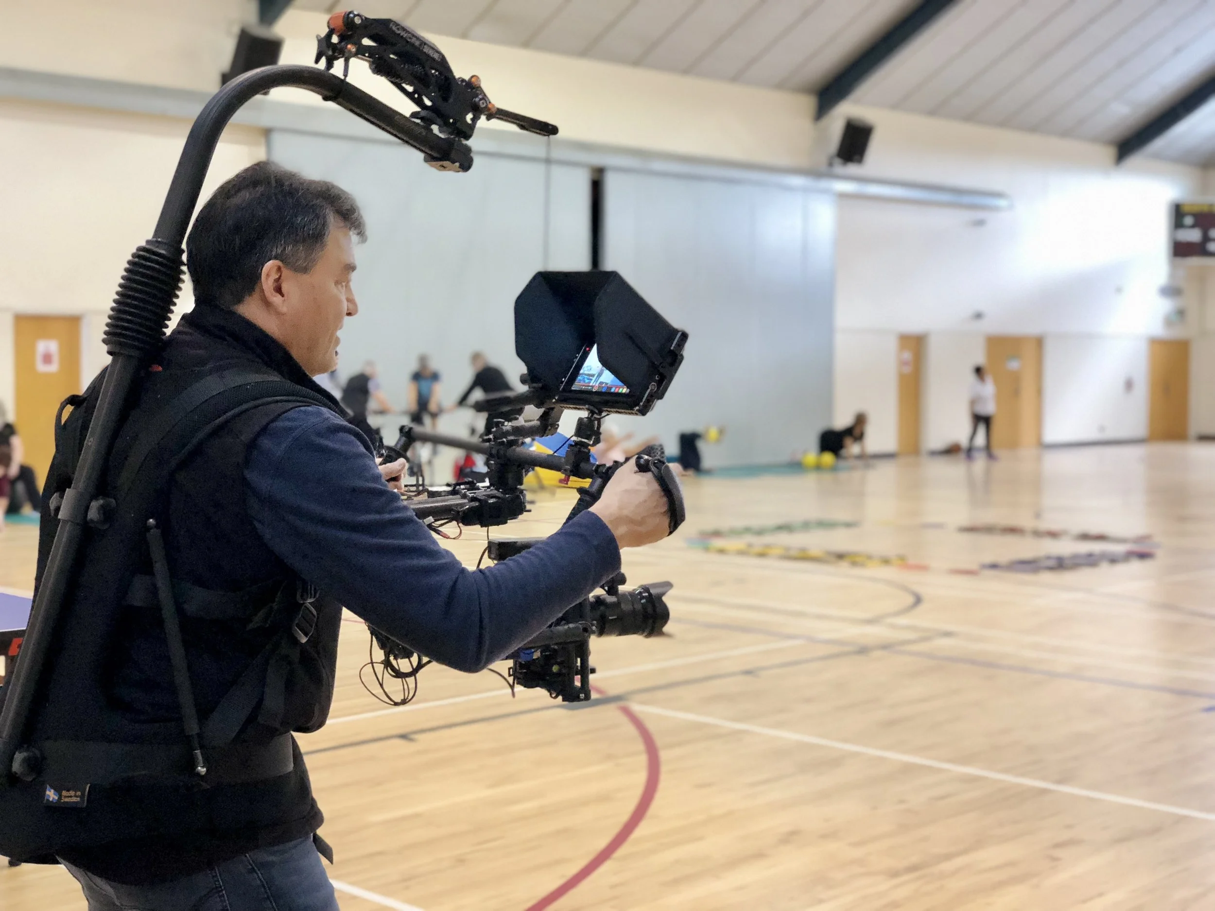 A man operating a professional video camera mounted on a shoulder rig inside a gymnasium, with people and activity in the background.