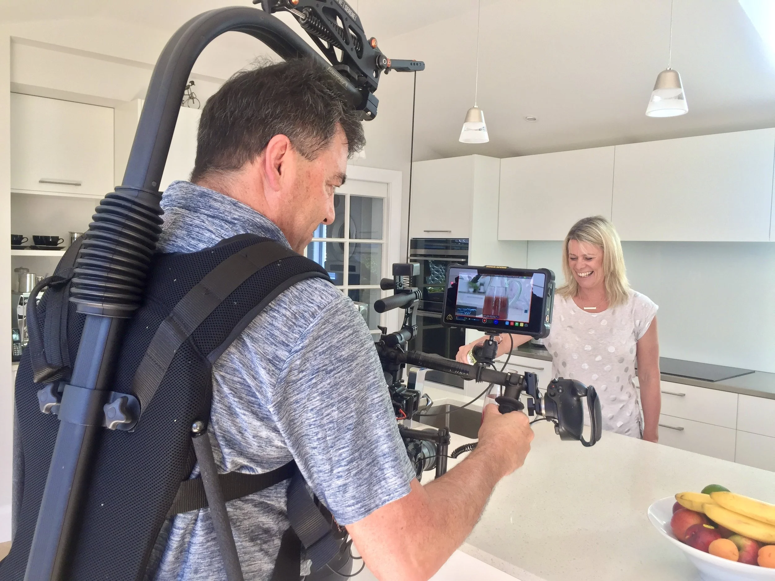 A man operating a camera rig film a woman in a white polka dot shirt, smiling in a modern kitchen with a fruit bowl on the counter.