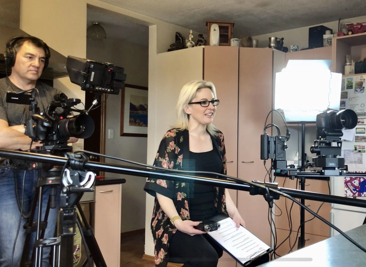 A woman with blonde hair and glasses is seated, holding a clipboard and a microphone, interviewing someones out of shot being filmed by a man with a large professional camera in a kitchen or dining area with cabinets and a bright window.
