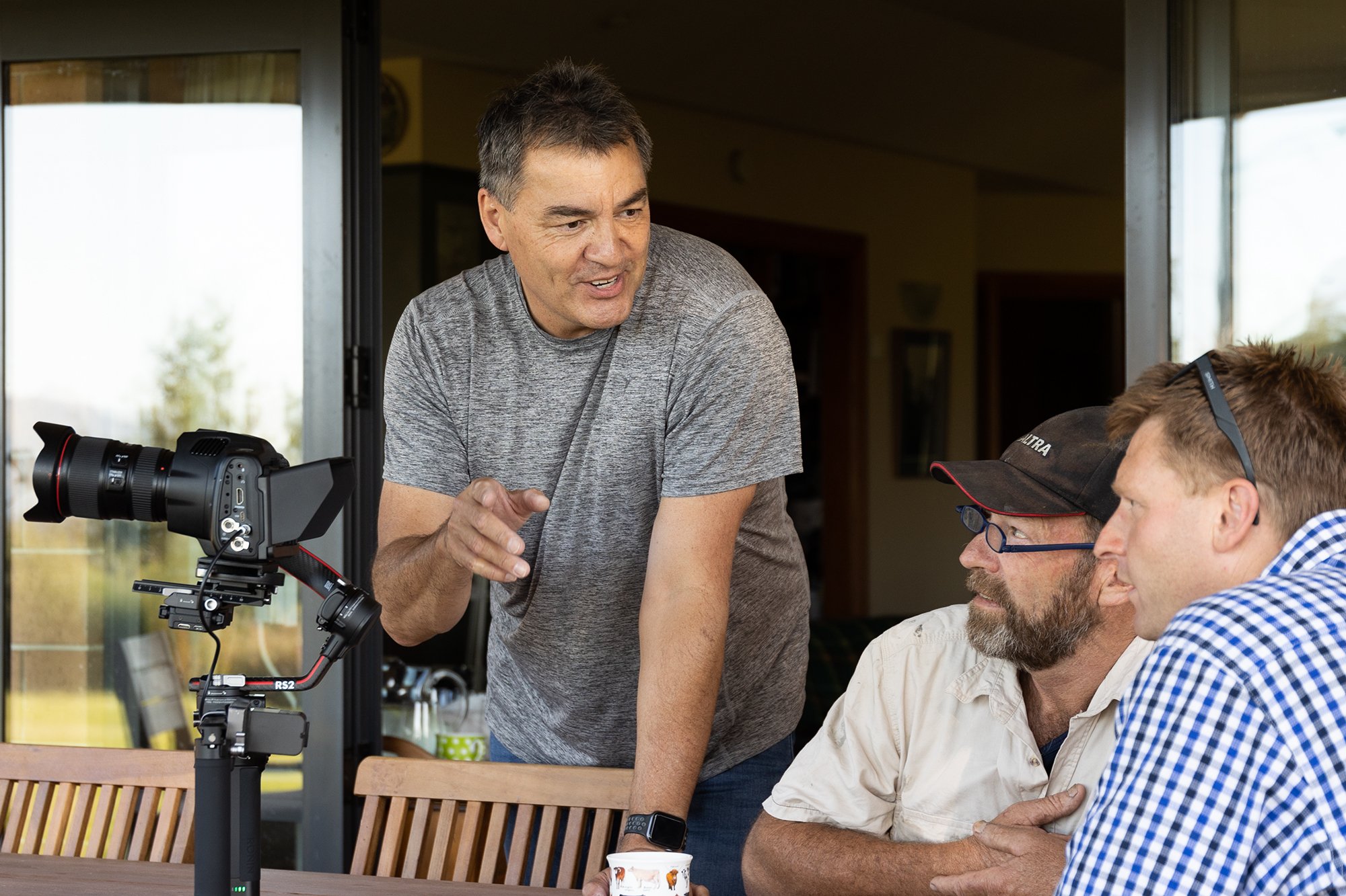 Three men are engaged in a discussion around a camera mounted on a tripod. The man standing is gesturing, while the two seated men listen attentively.