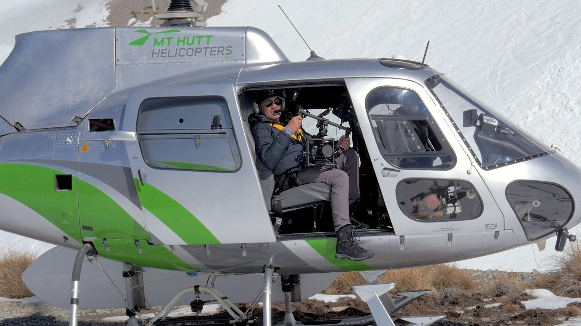 A man sitting inside a helicopter on a snowy terrain, holding a camera mounted on a stabilizer. The helicopter has the branding 'MT HUTT HELICOPTERS' on its side.