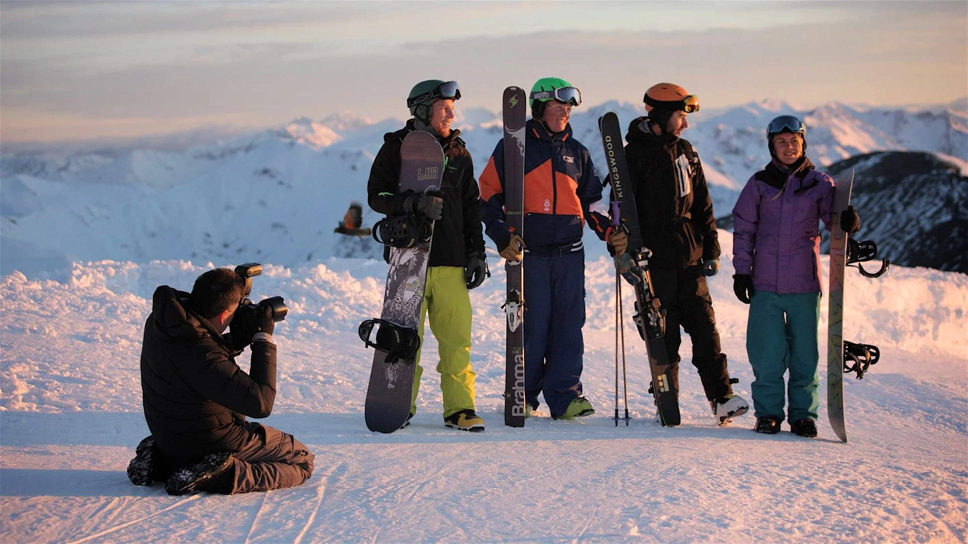 Group of five people in winter skiing gear holding skis, with a photographer taking their picture on snowy mountain landscape at sunset.
