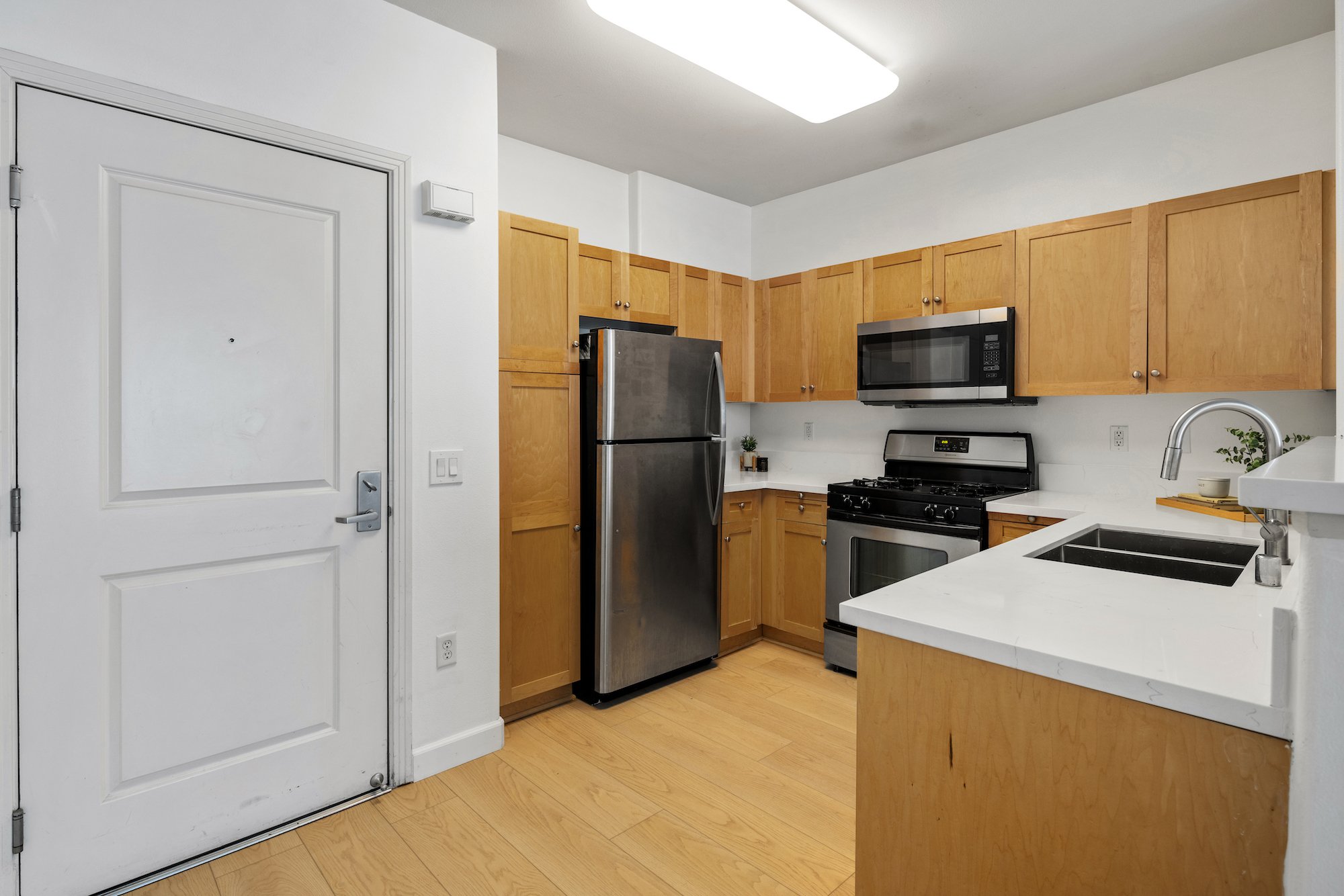 Kitchen with wooden cabinets, stainless steel refrigerator, stove, microwave, white countertops, and a white sink with a silver faucet.