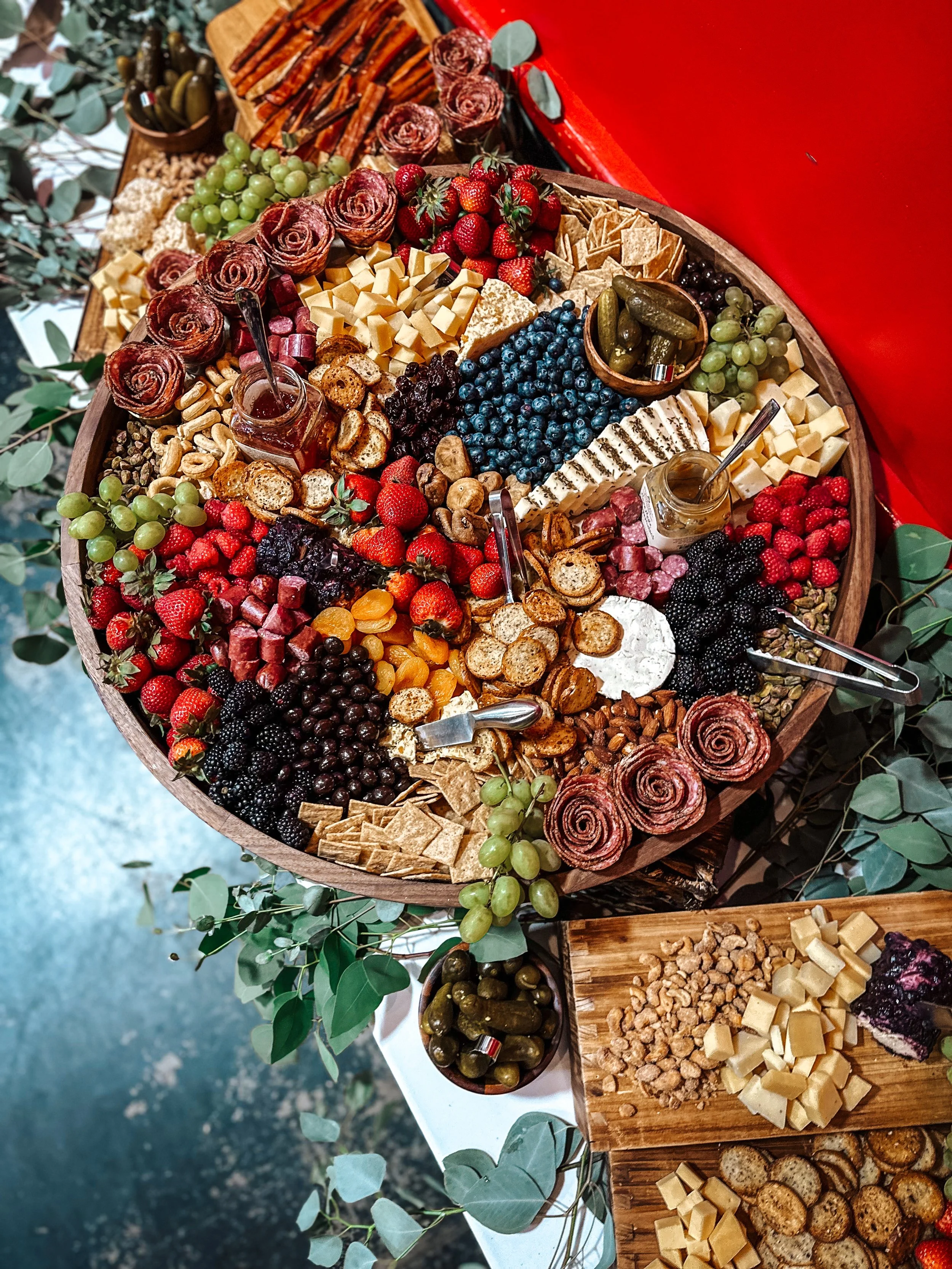 A large wooden platter filled with various cheeses, fruits, nuts, berries, and charcuterie items, surrounded by greenery and smaller wooden boards with additional accompaniments.