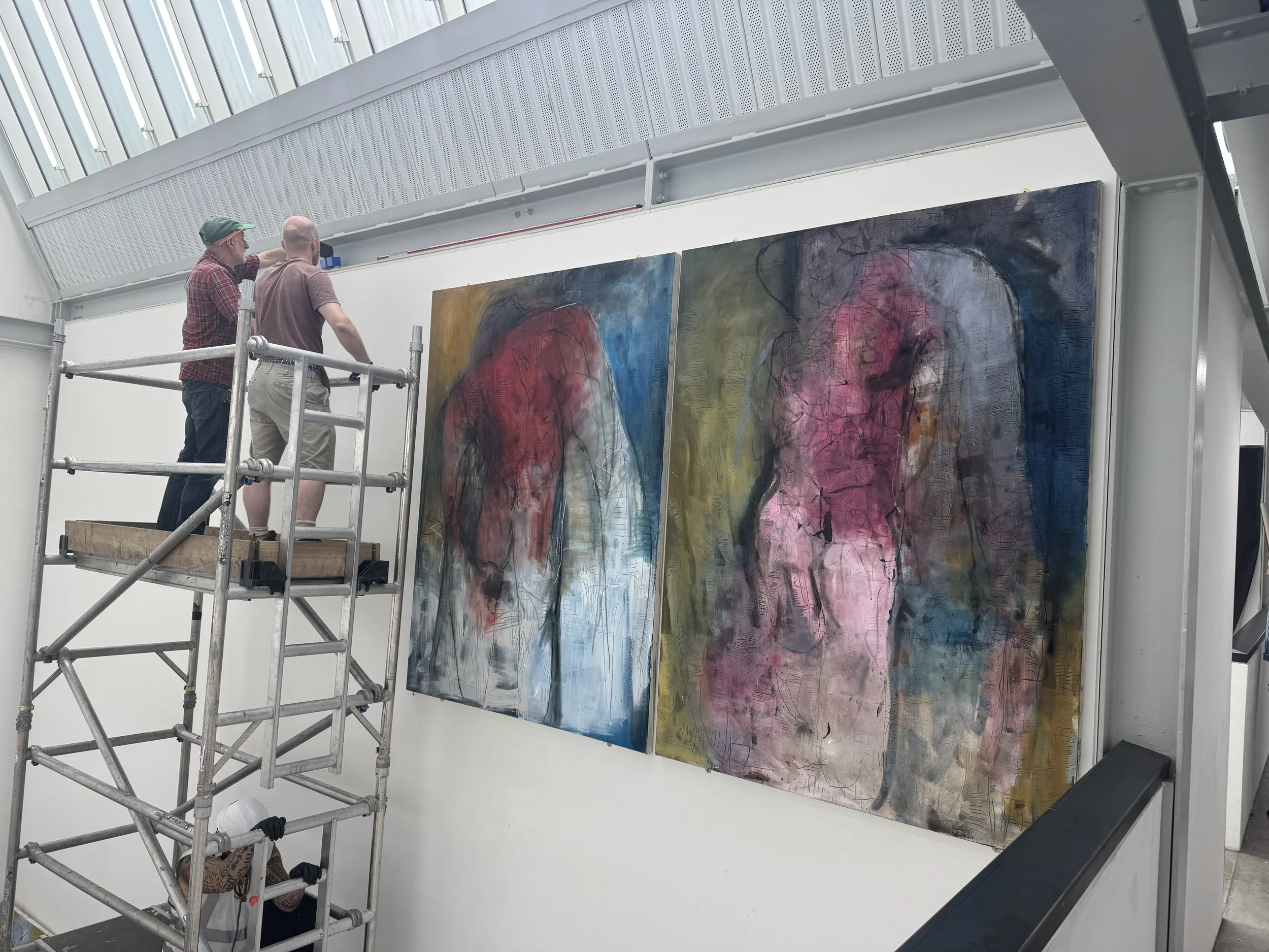 Two people are standing on a platform working on a large abstract painting at an art gallery. The painting features a humanoid figure with vivid colors and expressive lines.