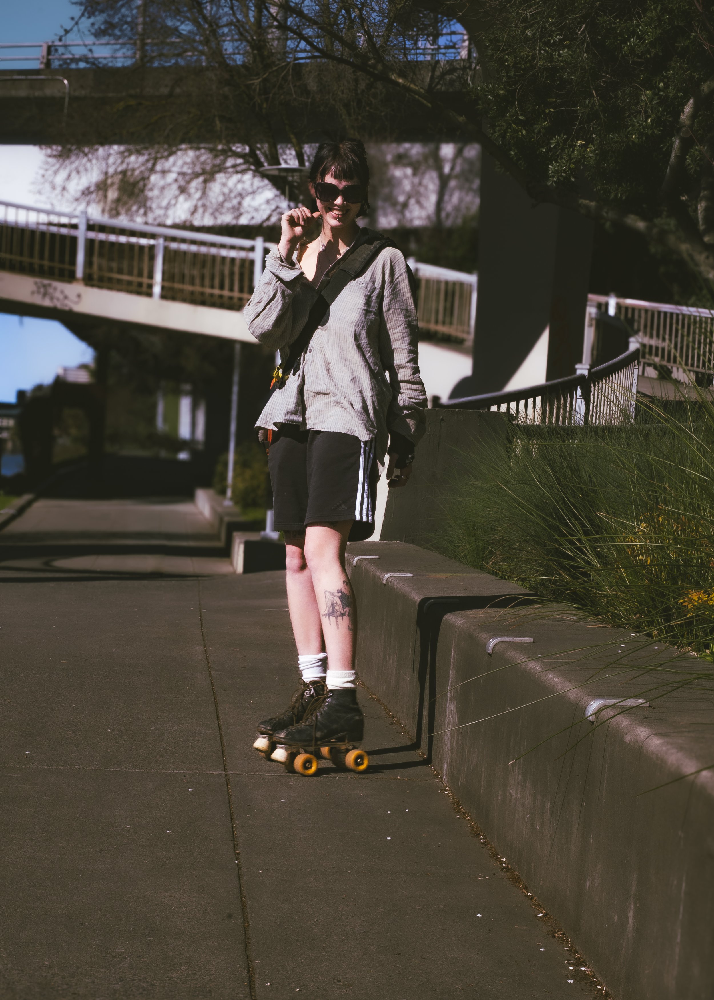 A woman wearing sunglasses, a light-colored shirt, and black shorts is roller skating and smiling outdoors near a concrete barrier, with trees and a sidewalk in the background.