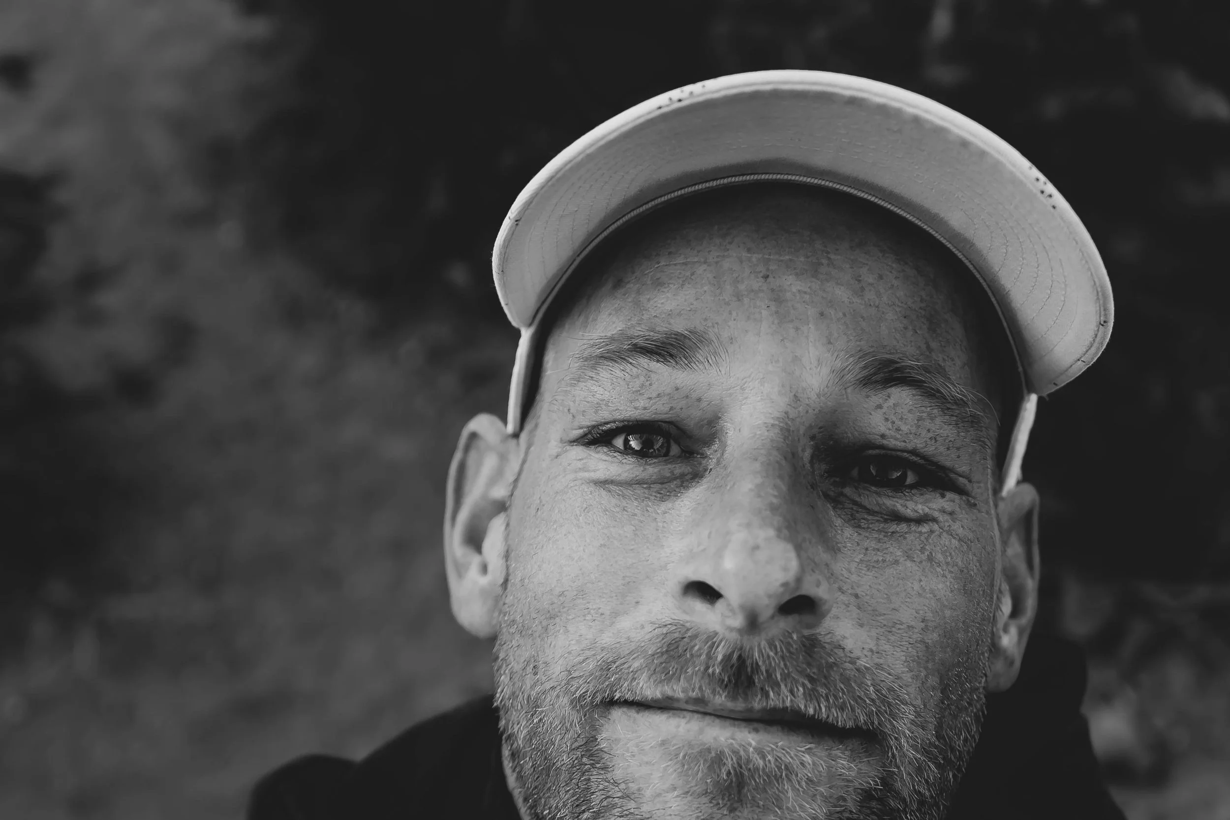 Close-up black and white photograph of a man's face with a hat, showing his eye, part of his nose, and a beard.