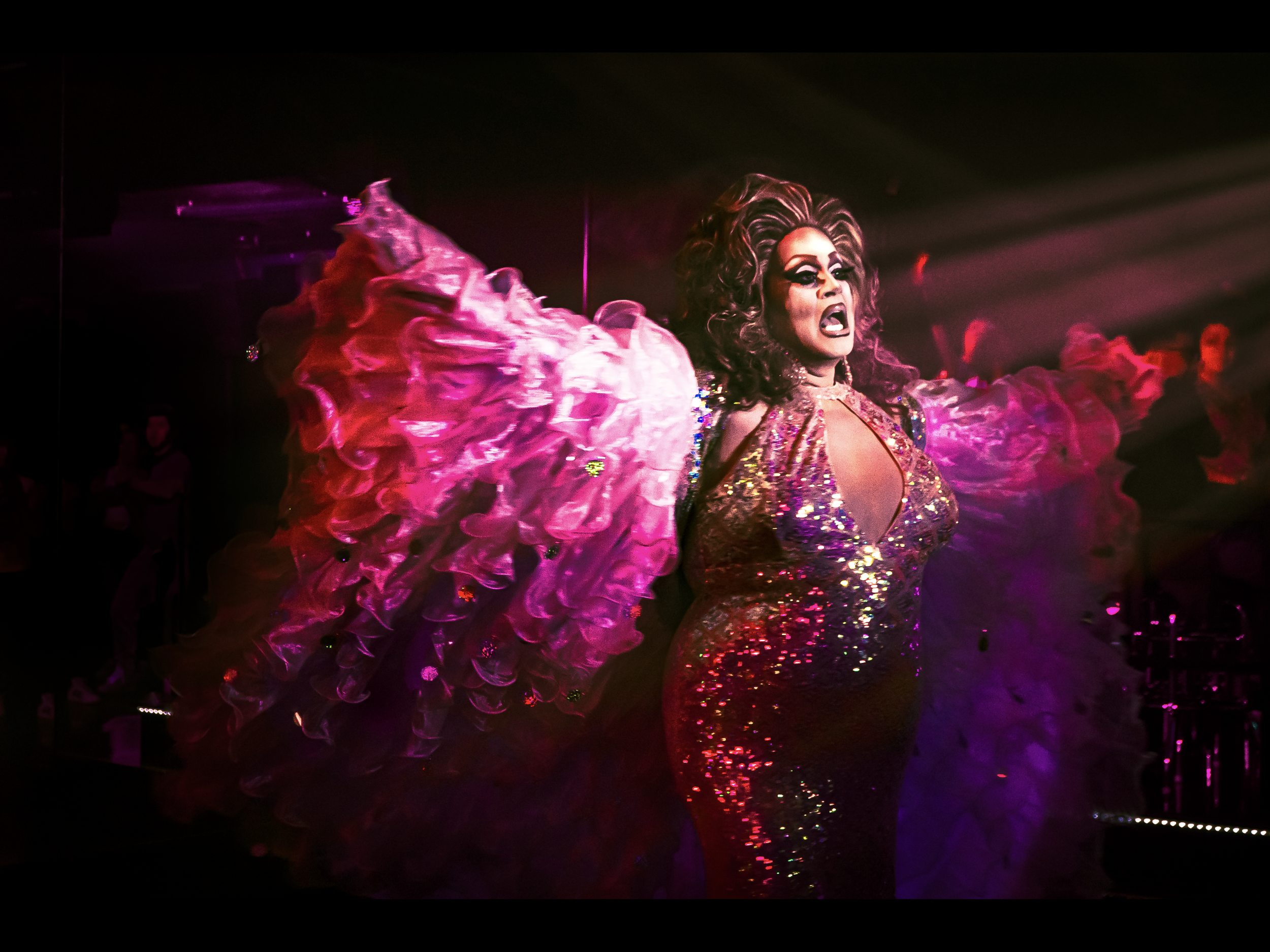 A drag performer with bold makeup, voluminous hair, and a sequined dress, appears to be on stage in a nightclub with pink and purple lighting, surrounded by an audience.