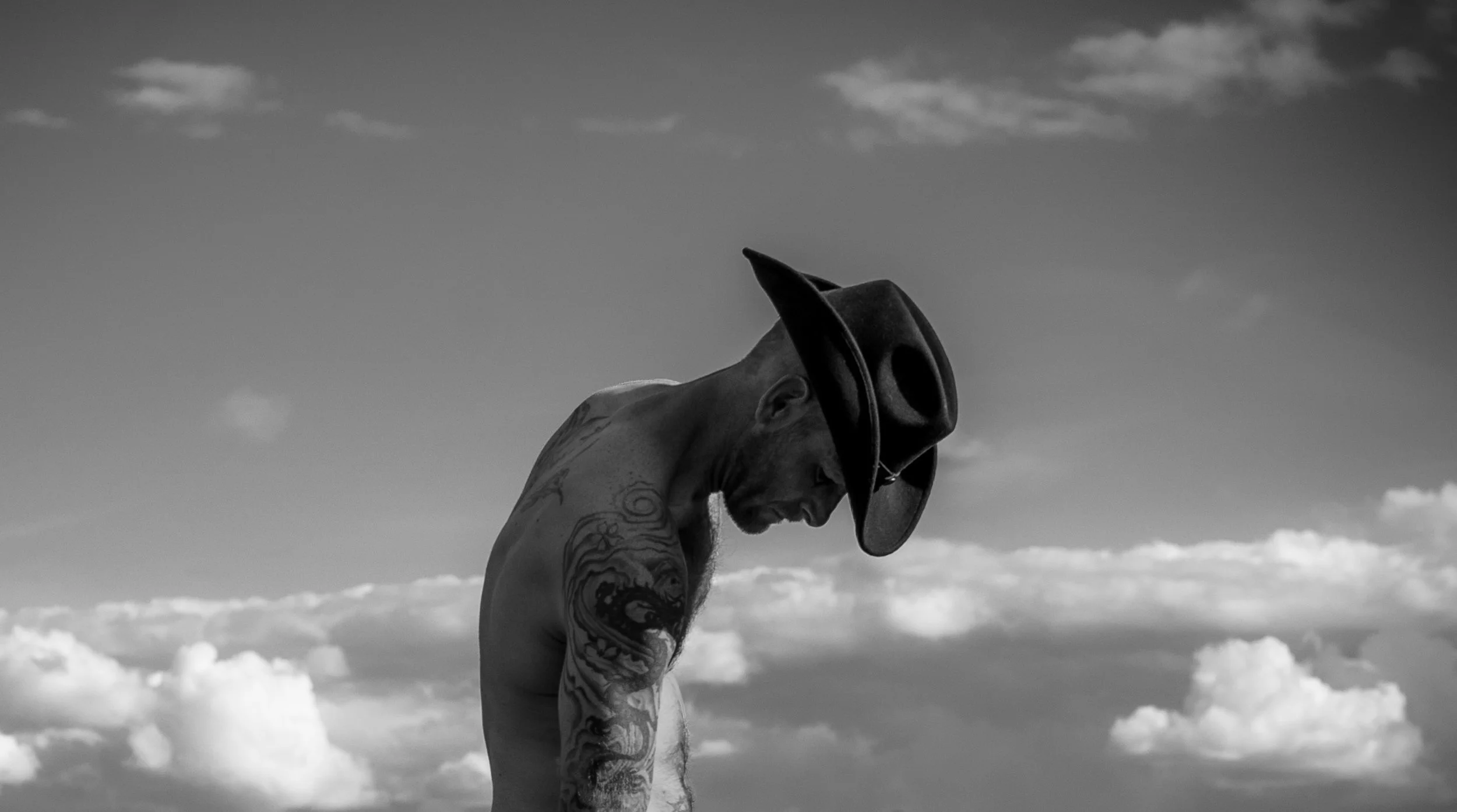 Black and white photo of a shirtless man with tattoos wearing a cowboy hat, looking down with a cloudy sky in the background.