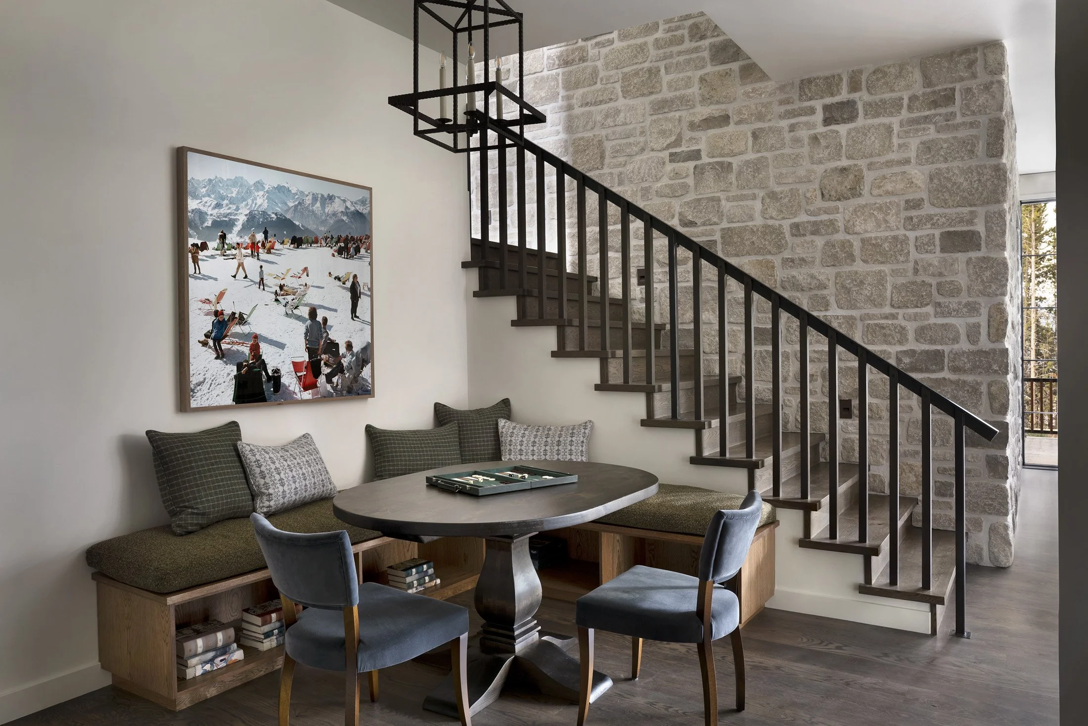 Rustic modern luxury ski home in Big Sky Montana with a corner banquette game table next to a staircase with exposed stone walls, iron railings, iron chandelier, and funishings inspired by colors of nature designed by Hive Home