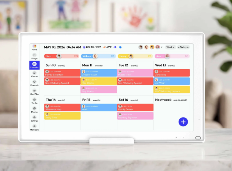 Digital tablet displaying a weekly family calendar with scheduled activities in various colors, including breakfast, work, therapy, swimming lessons, and family dinner, on a white marble surface with blurred children's artwork in the background.