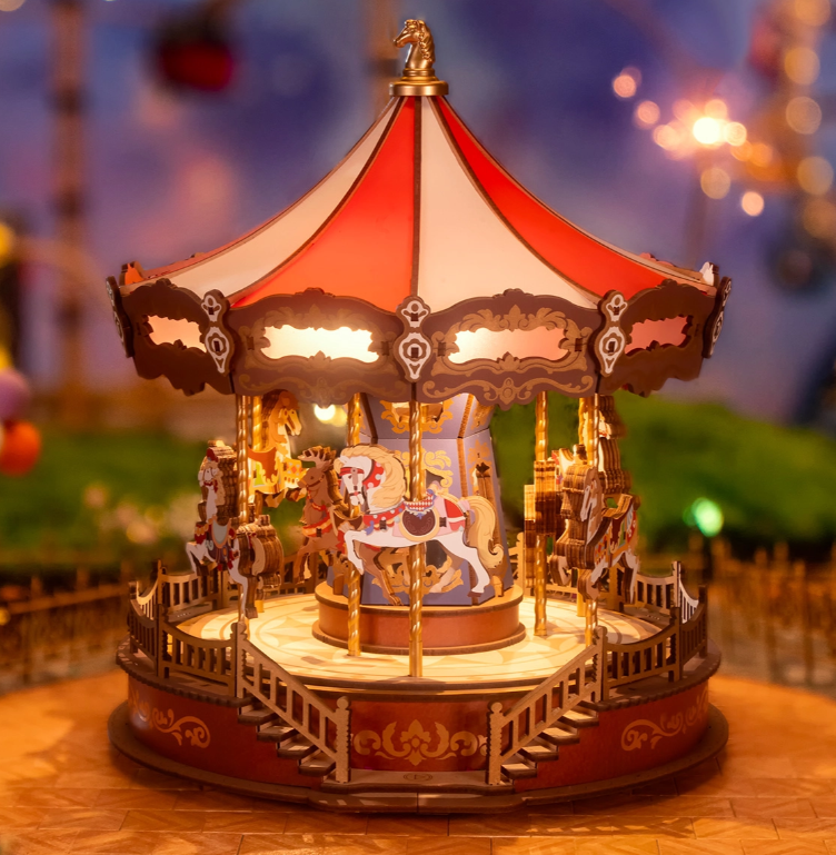 A detailed miniature carousel with a red and white canopy, ornate horses with saddles, and a surrounding fence, set against a blurred background with warm lighting.