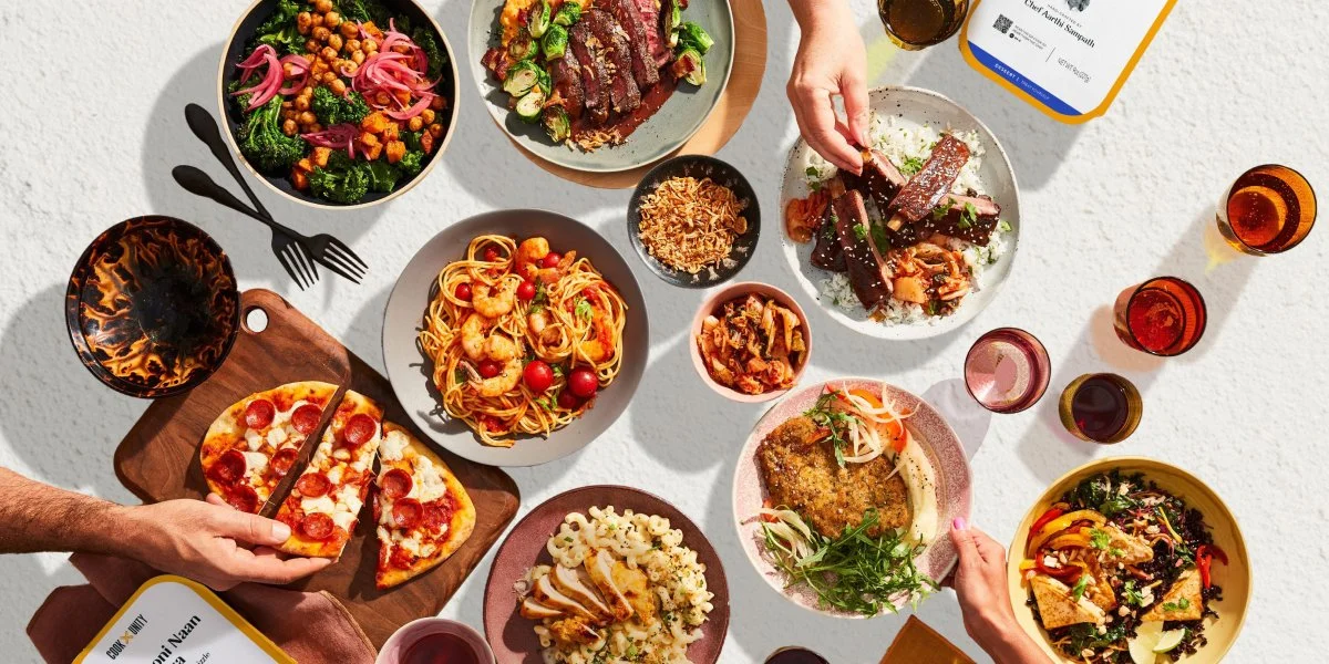 A table filled with various dishes including a vegetable salad, a plate of sliced steak, a bowl of spaghetti with shrimp and cherry tomatoes, a pizza with pepperoni, a fried cutlet with vegetables, and a mixed salad. There are also four drinks along the side.