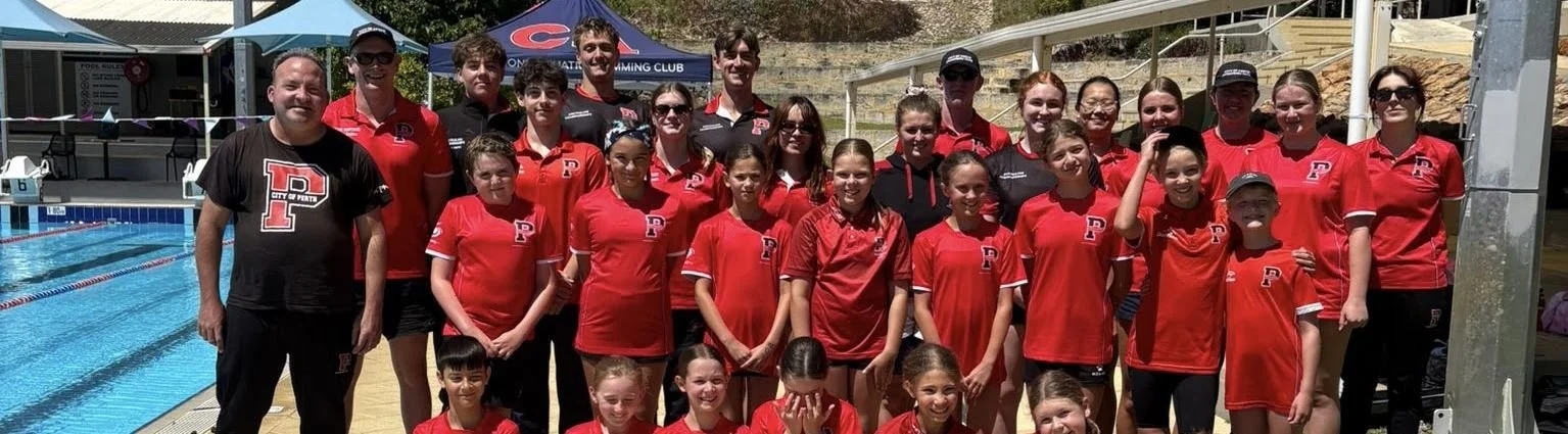 Group of young swimmers and coaches at an outdoor swimming pool, wearing red team uniforms, posing for a photo.