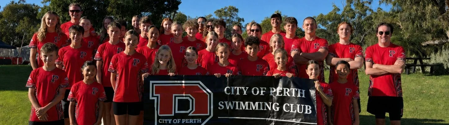 Group photo of young swimmers and coaches from the City of Perth Swimming Club, all wearing matching red shirts, standing outdoors on a grassy area with trees in the background, holding a banner that reads 'City of Perth Swimming Club'.
