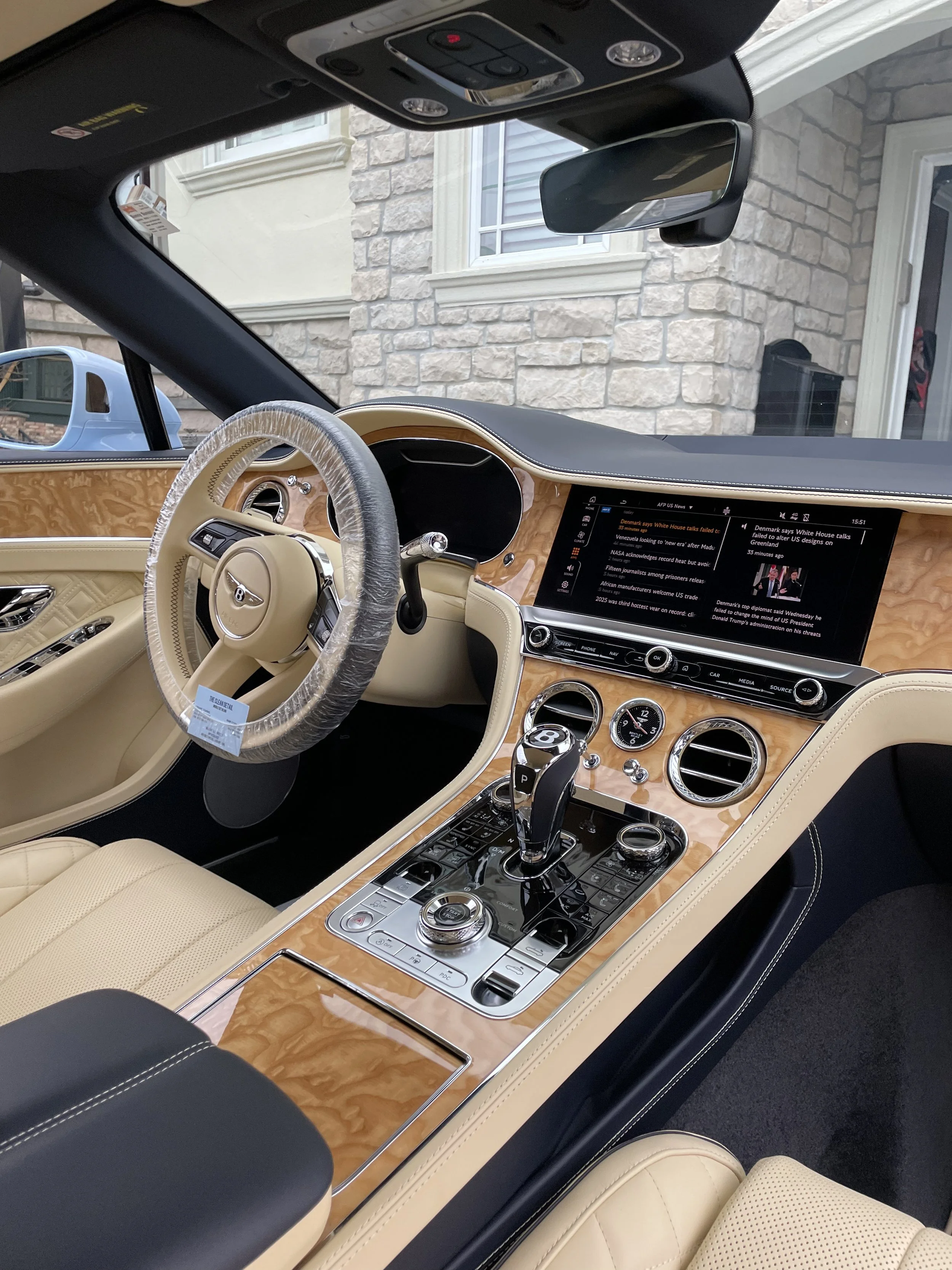 Interior of a luxury car with tan and black leather seats and a woodgrain dashboard. The steering wheel is wrapped in plastic. The center console has a gear shifter, multimedia controls, and a digital screen displaying news.