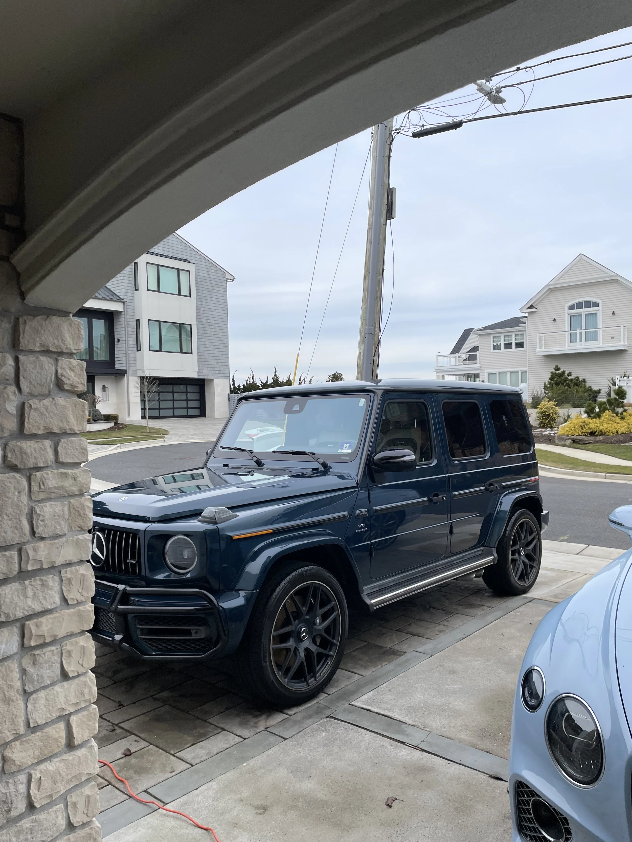 A blue Mercedes-Benz G-Class SUV parked on a driveway in front of a modern house, with another white car partially visible in the foreground.