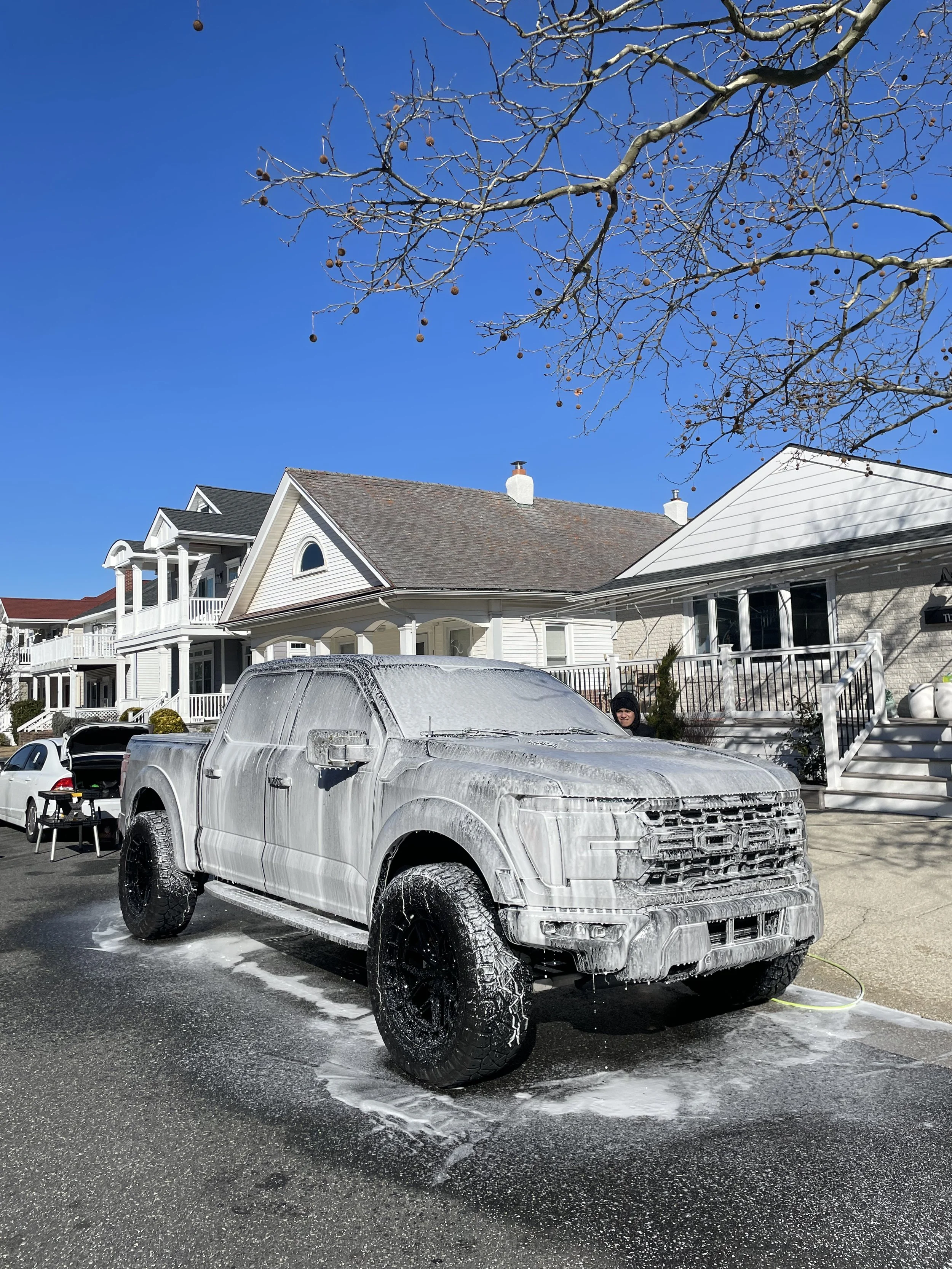 A large truck covered in soap foam during a car wash on a residential street on a sunny day with a clear blue sky.
