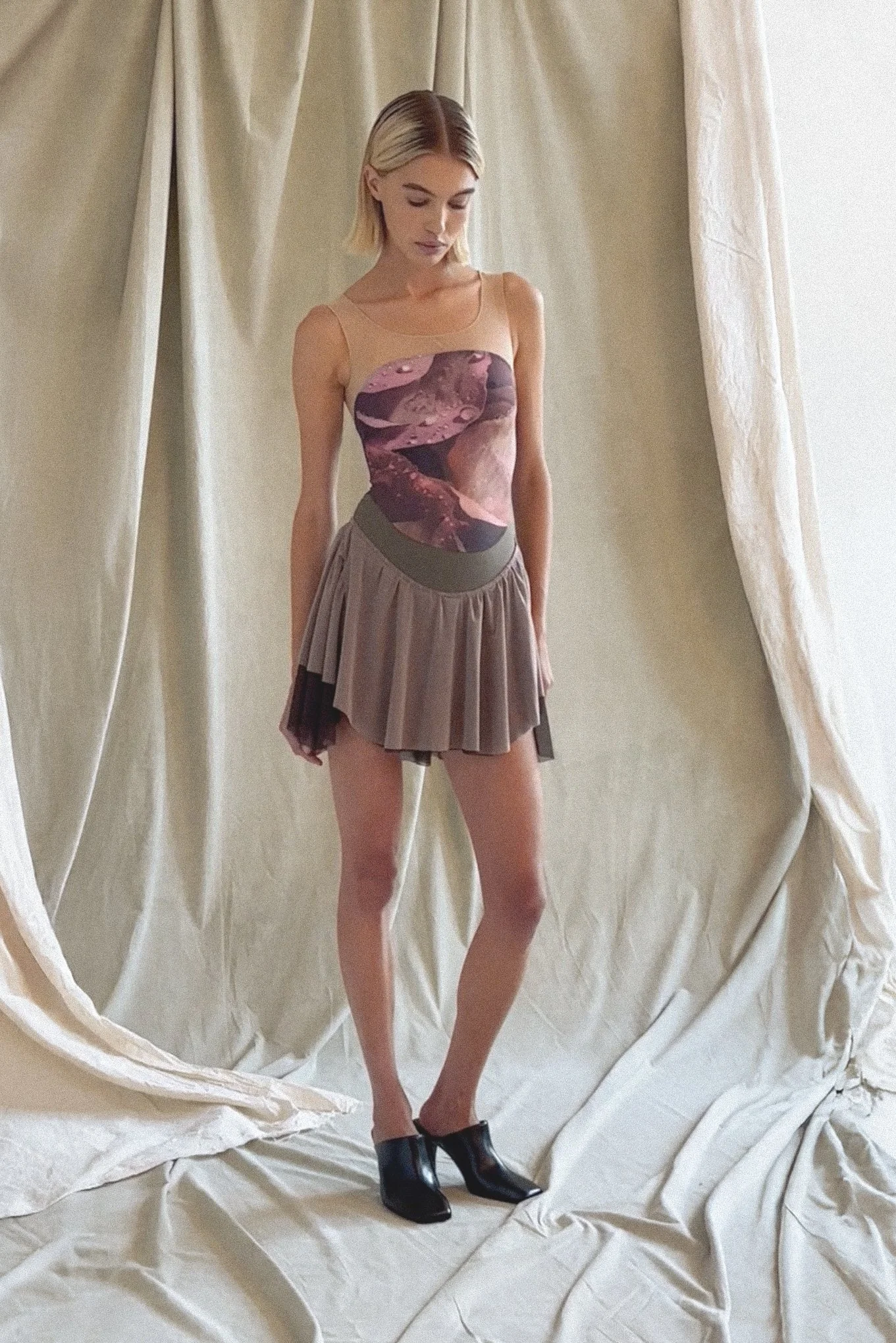 A young woman with blonde hair wearing a sleeveless top with an abstract print, a pleated mini skirt, and black ankle boots standing in front of beige curtains.