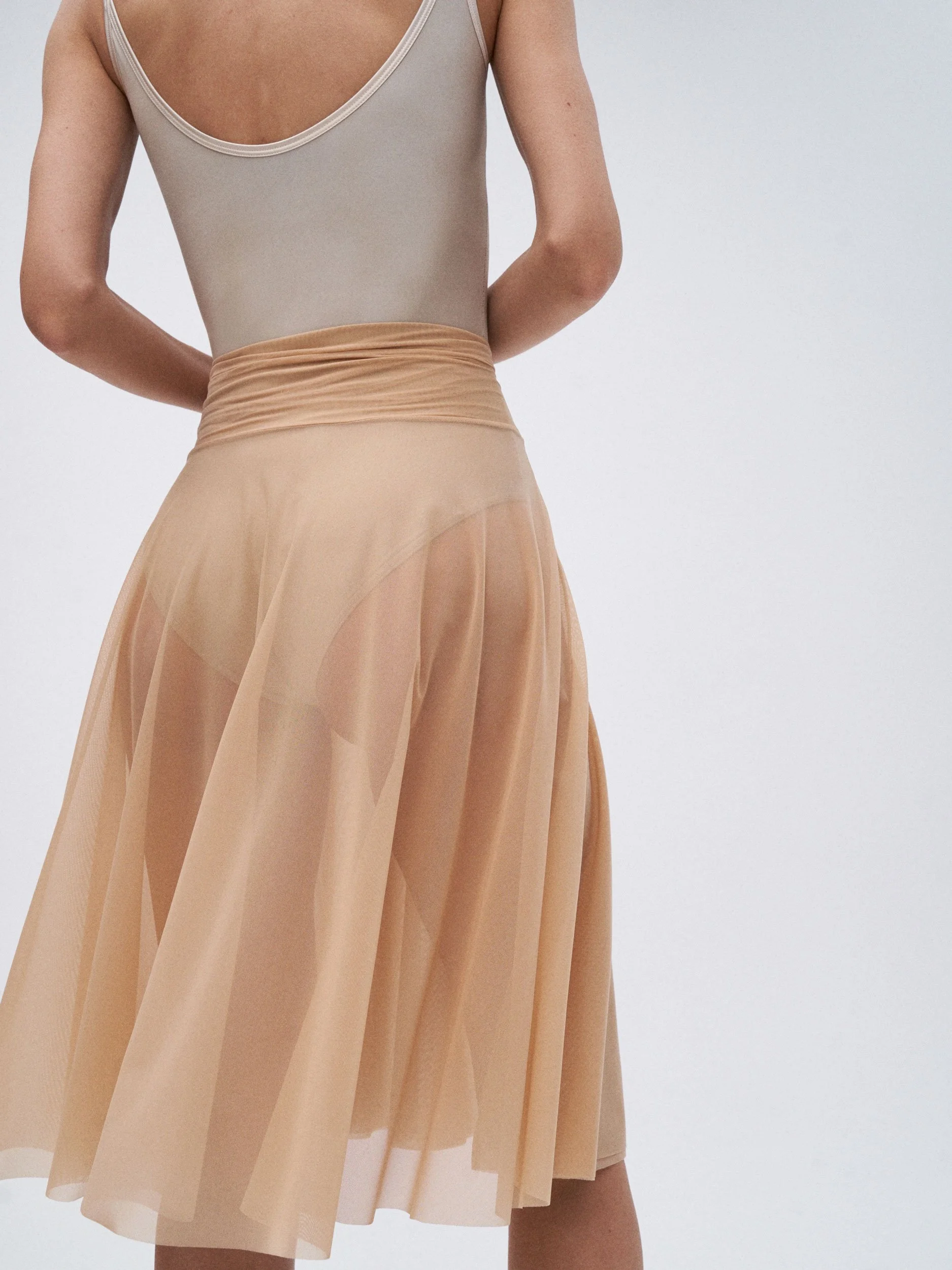 A woman wearing a nude-colored bodysuit and a sheer peach-colored midi skirt, with her arms behind her back, against a plain white background.