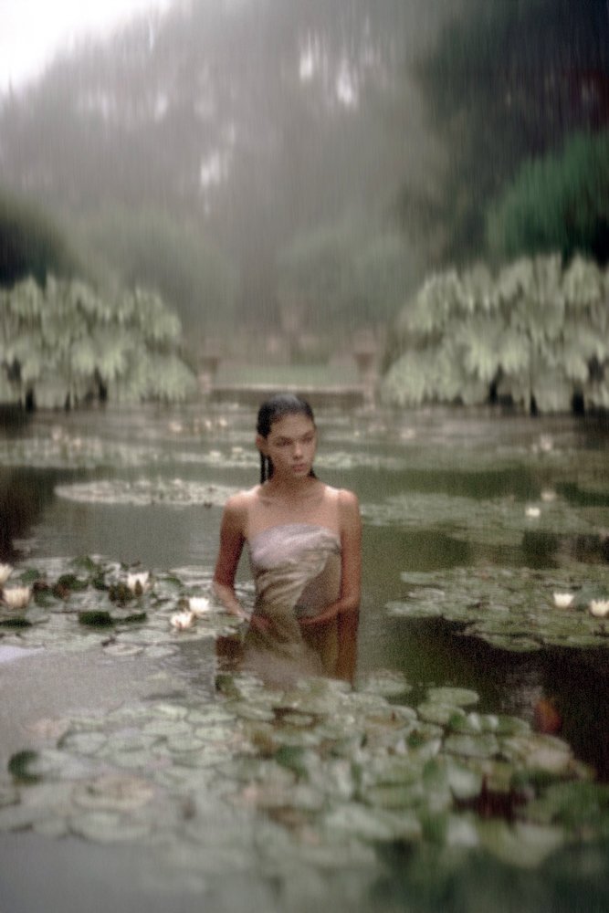 A young woman stands in a pond surrounded by water lilies, with a misty, lush greenery background.