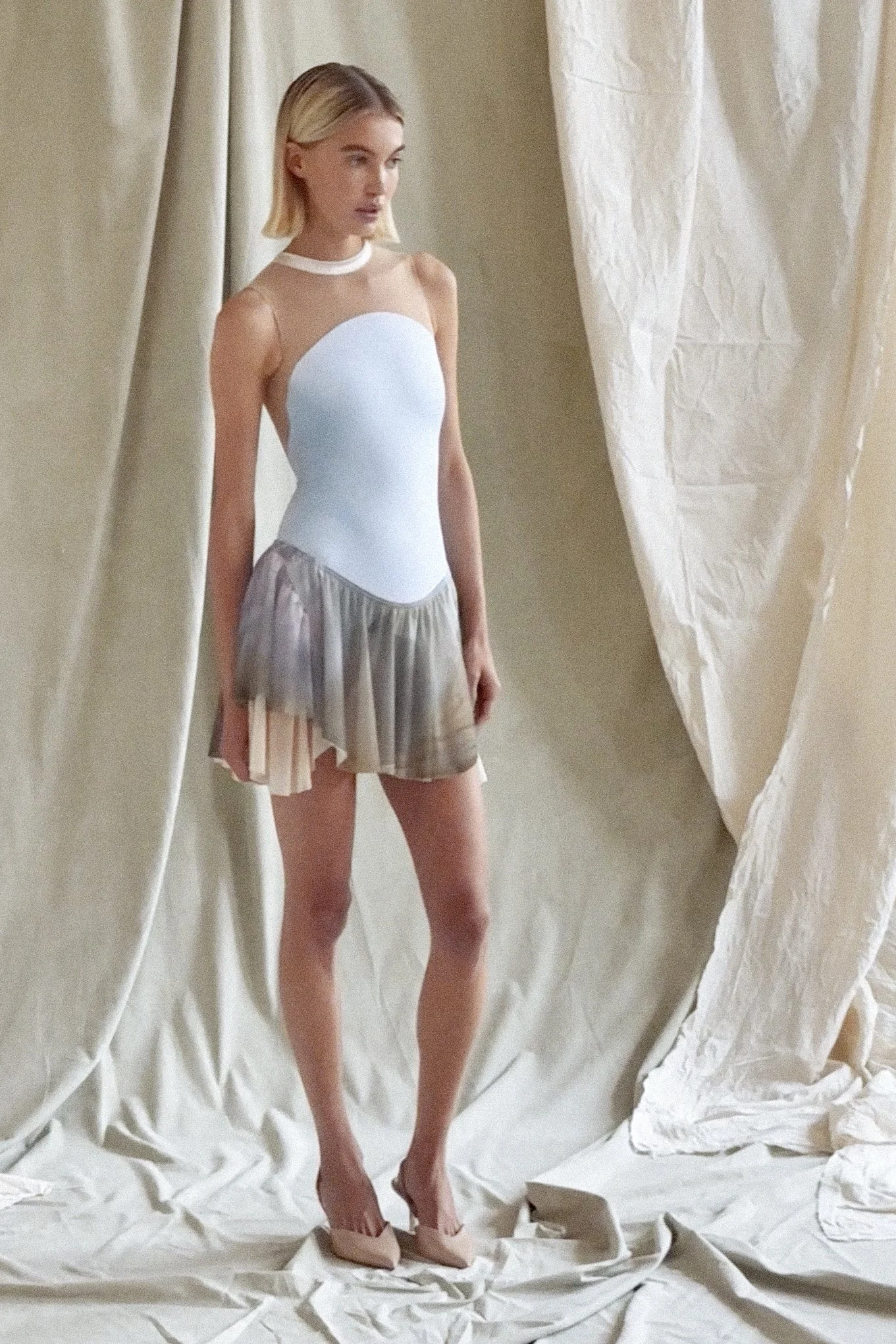 A woman with short blonde hair standing on a beige fabric floor surrounded by beige curtains.