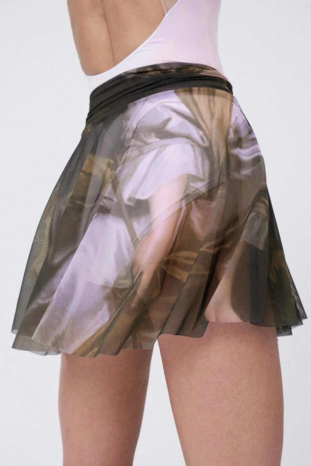 Close-up of a woman wearing a sheer pleated skirt with a printed pattern and a sleeveless top, with a plain white background.