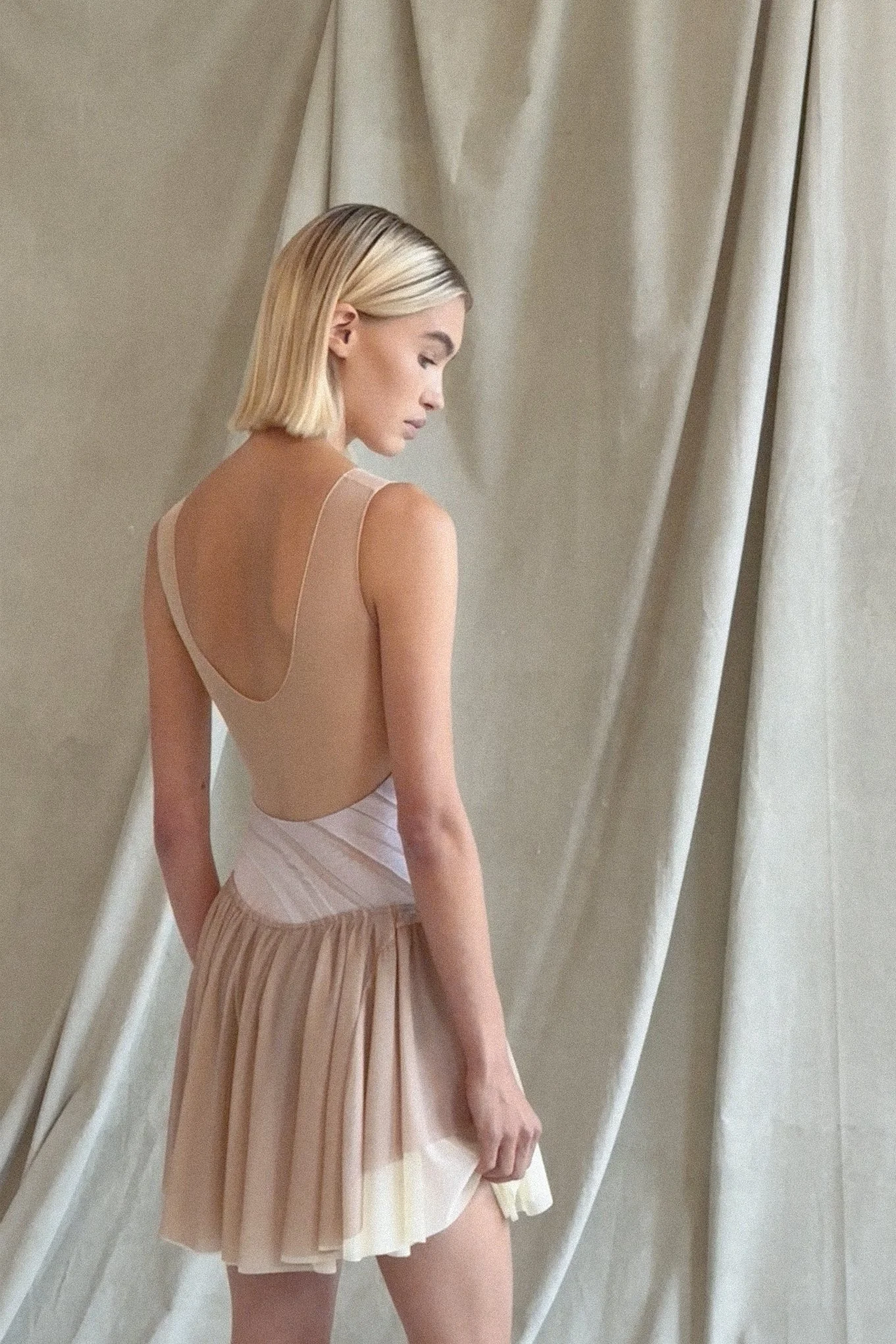 A woman in a beige dress standing near cream-colored curtains, looking down with her hair styled in a short blonde bob.