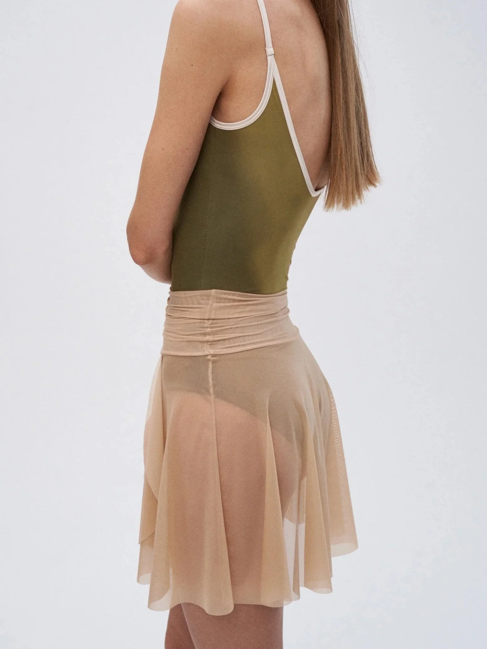 A woman in an olive green sleeveless top with white trim and a sheer beige skirt against a plain white background.
